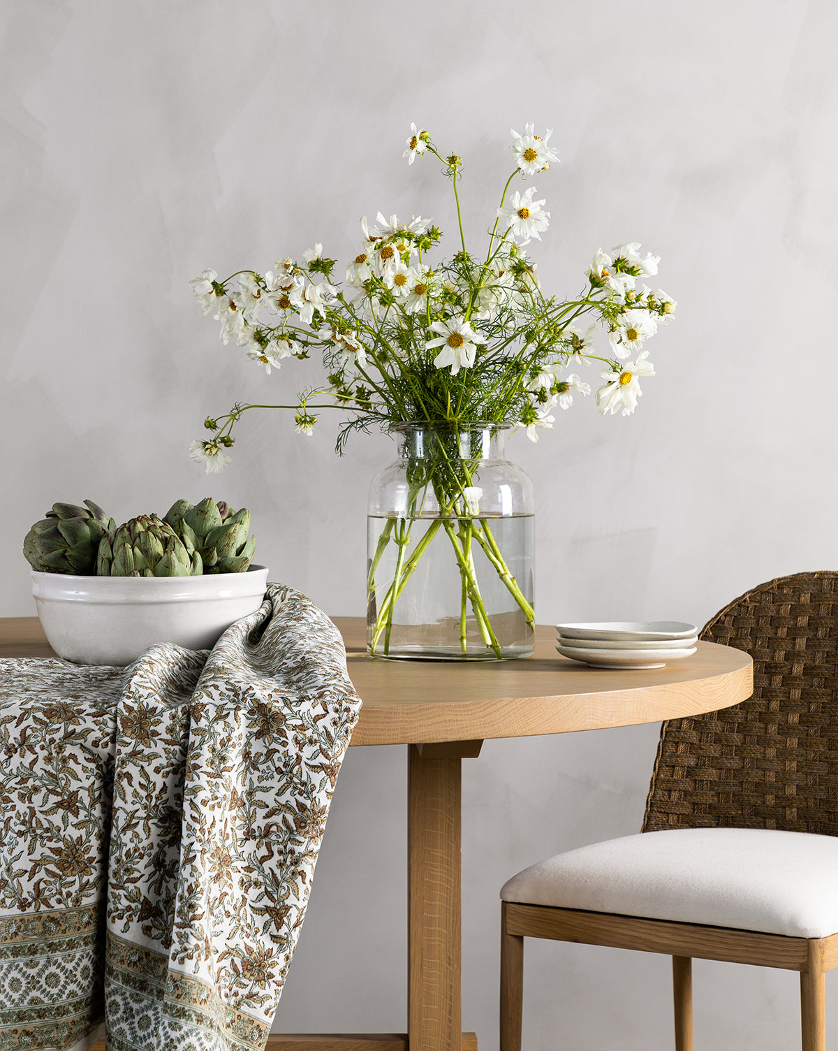 A glass vase of white daisies sits on a round wooden table next to the Kern Glass Jar, a bowl of artichokes, two stacked plates, and a patterned cloth. A woven chair is partially visible on the right.