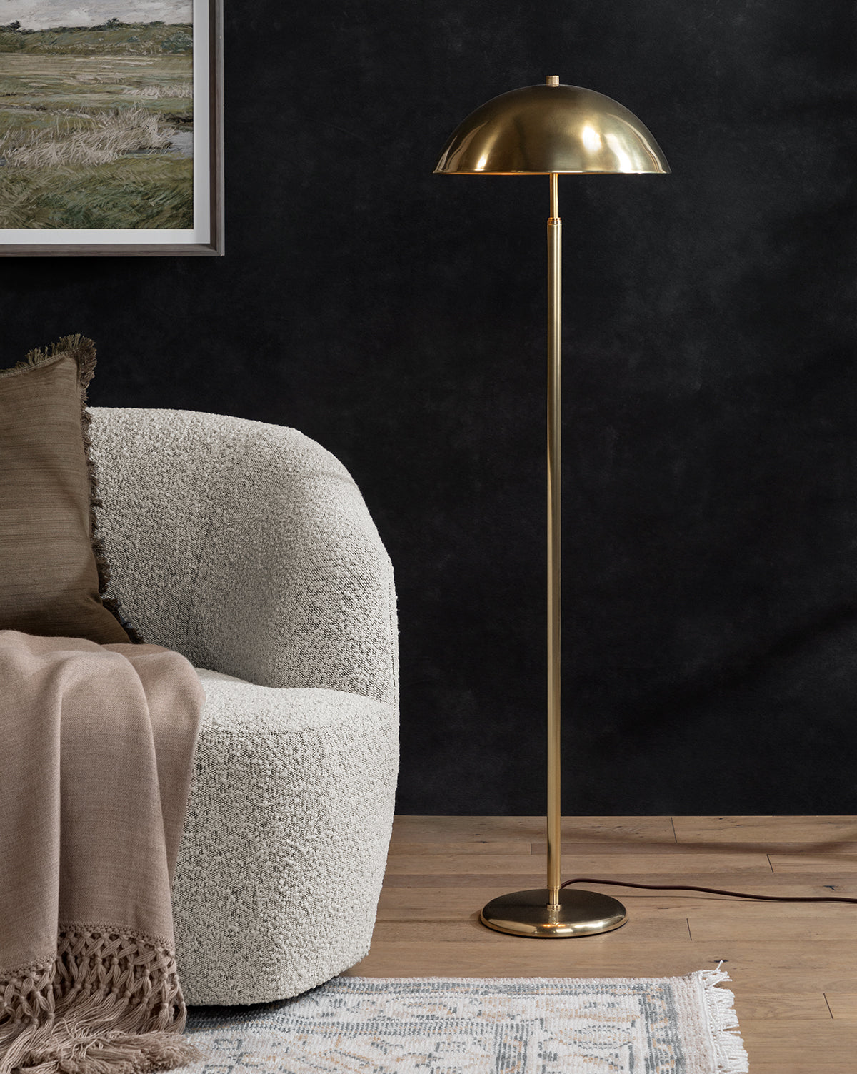 A modern living room with a textured white armchair, beige throw blanket, the Aldorno Floor Lamp featuring a dome-shaped shade, a blue patterned rug, and a framed landscape painting on a dark wall, by McGee & Co.