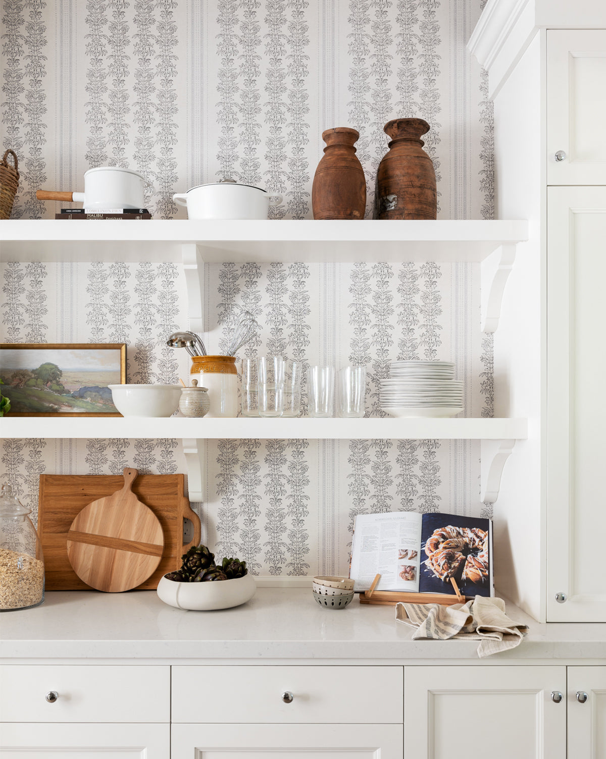 Open kitchen shelves display glasses, bowls, cutting boards, and jars against York Wallcoverings Demi Floral Stripe Wallpaper. On the white countertop below, an open cookbook, cups, and a cloth are arranged—McGee & Co.
