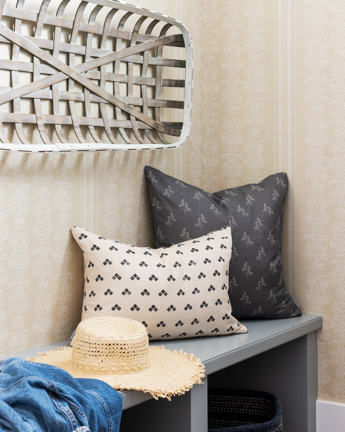 A straw hat and blue denim jacket rest on a gray bench with two patterned pillows, set against York Wallcoverings’ Demi Floral Stripe Wallpaper. Above, a woven basket tray adds extra charm with subtle floral motifs—McGee & Co.