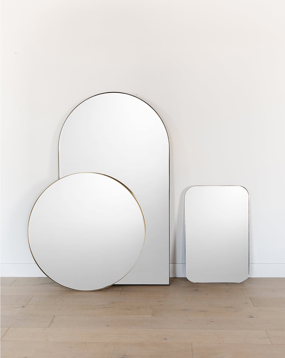 Three modern mirrors with thin frames stand against a white wall on a wooden floor: one tall arch-shaped entryway mirror, the Jace Inset Circle Mirror, and a smaller rectangular mirror by McGee & Co.