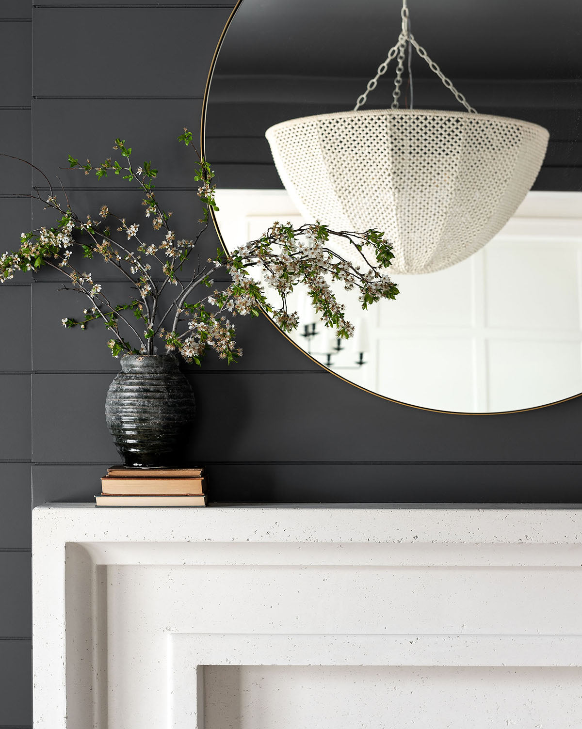 A black vase with flowering branches rests on books atop a white mantel, below the Jace Inset Circle Mirror, which reflects a textured white chandelier against a dark paneled wall, by McGee & Co.