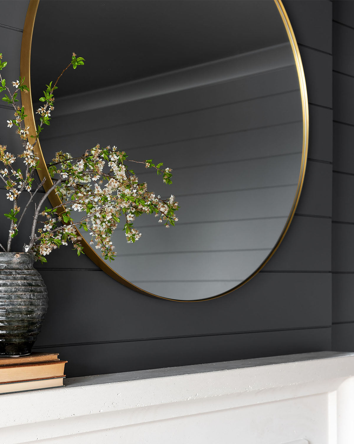 The Jace Inset Circle Mirror with a gold frame hangs on a dark shiplap wall above a white mantel displaying stacked books and a ceramic vase with blooming branches, all reflected in the mirror’s shatterproof glass by McGee & Co.