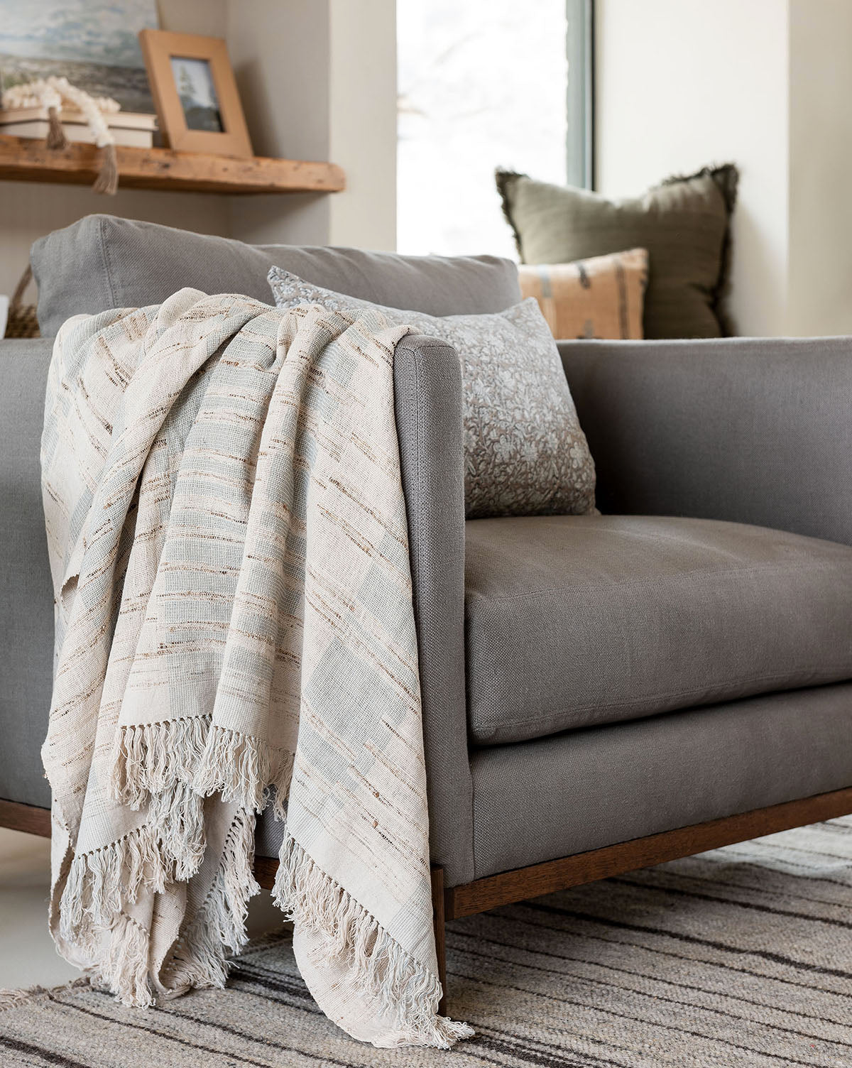 A gray armchair with a wooden base sits in a cozy room, draped with the Theodora Block Stripe Throw and accented by a patterned pillow. A striped rug, cushions, and decor items add warmth to the inviting setting.