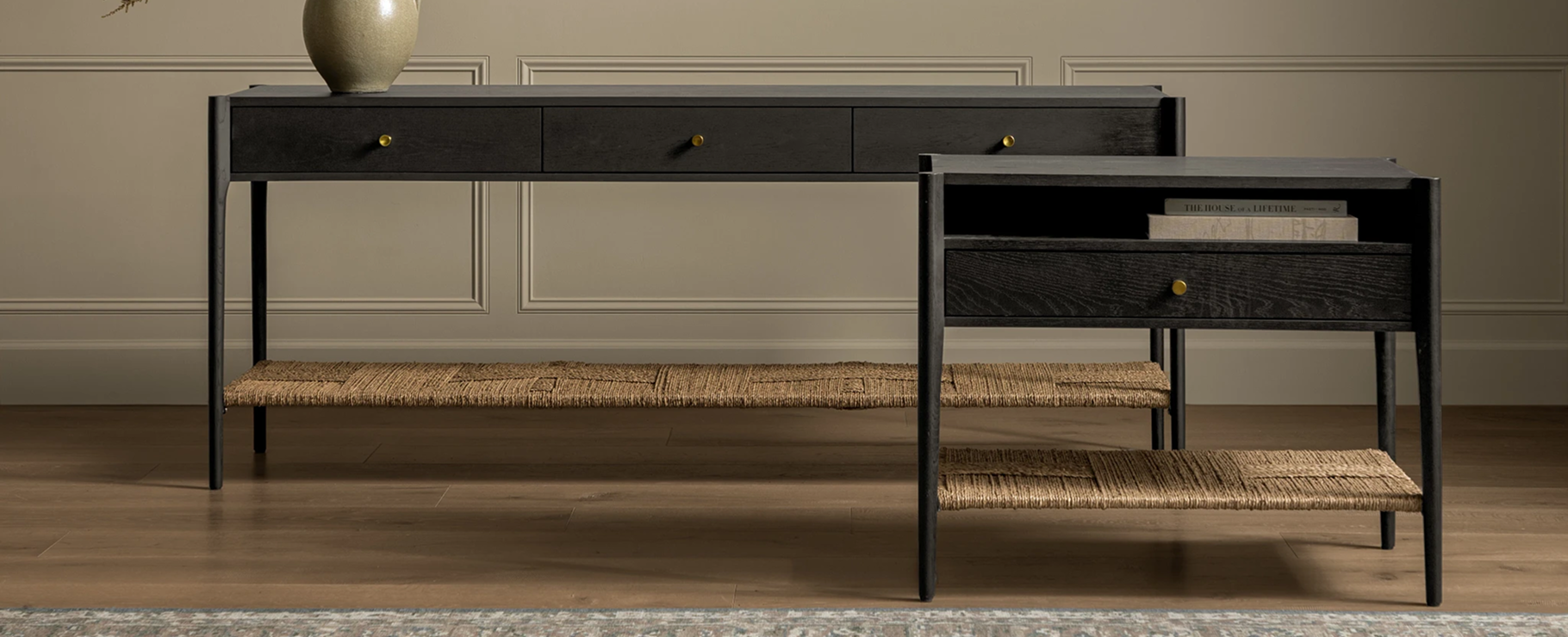 A black wood console table and matching side table with woven lower shelves and gold knobs stand on a wooden floor against a beige, paneled wall, by McGee & Co. A vase decorates the console table.