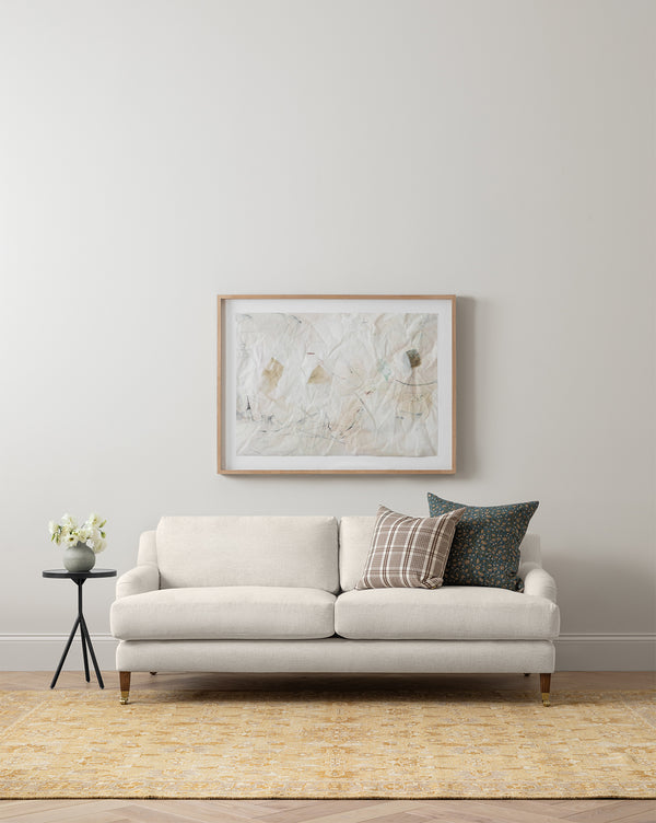 A modern living room with the customizable Lucille English Roll Arm Sofa 90.5 by Community in beige, down-wrapped cushions, two decorative pillows, a round side table with a flower vase, abstract art on a light wall, and a yellow rug on wood floors by McGee & Co.