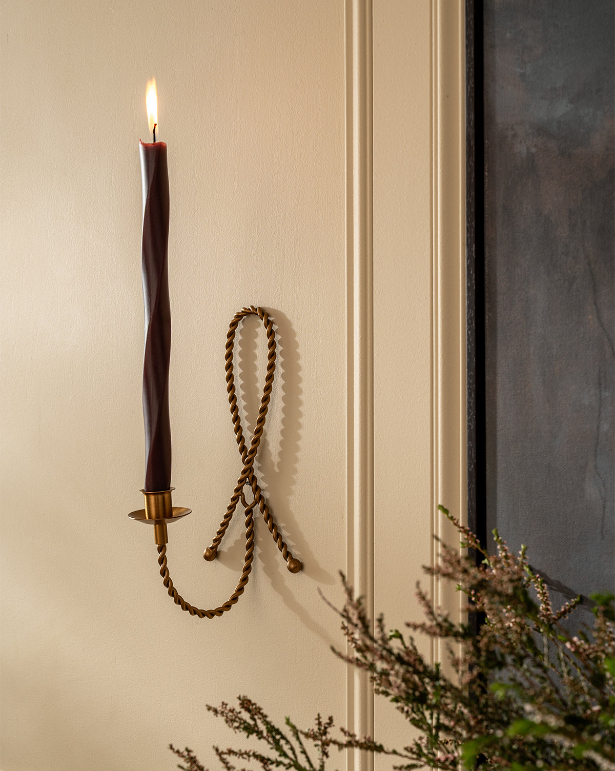 A lit twisted burgundy candle is placed in the Hearst Taper Holder, a sculptural brass piece with a decorative loop, mounted on a cream wall. Greenery appears in the bottom right corner, by McGee & Co.