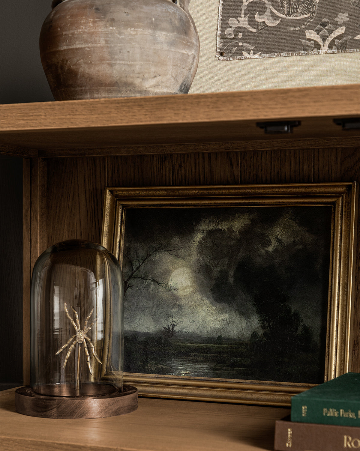 A wooden shelf showcases Storm on the Lake, a gold wood-framed painting of a spooky landscape under a moon, next to a large clay pot, a glass dome with a star-shaped object, and two stacked green books—perfect for unique home decor.