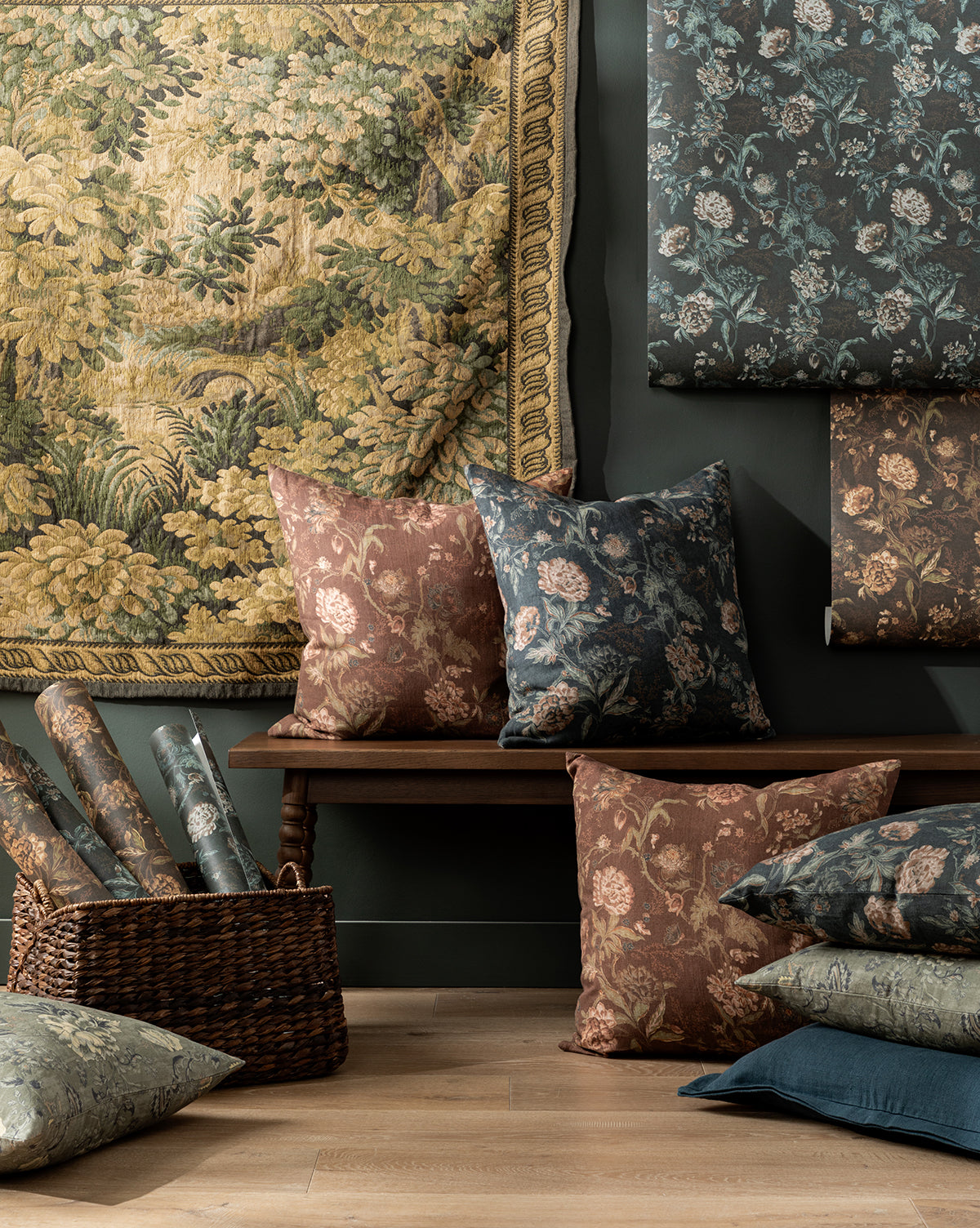 A cozy interior with painterly floral tapestries, blue and brown patterned cushions on a wooden bench, Hestercombe Floral Wallpaper by Makerspalm in a wicker basket, all set against a dark wall. McGee & Co.