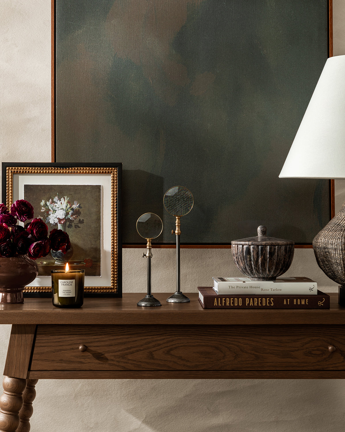 The Heathcliff Console with a walnut oak finish displays books, a lidded bowl, lamp, two magnifying glasses, a candle, dark red flowers in a vase, and a framed floral painting beneath a large abstract artwork by McGee & Co.