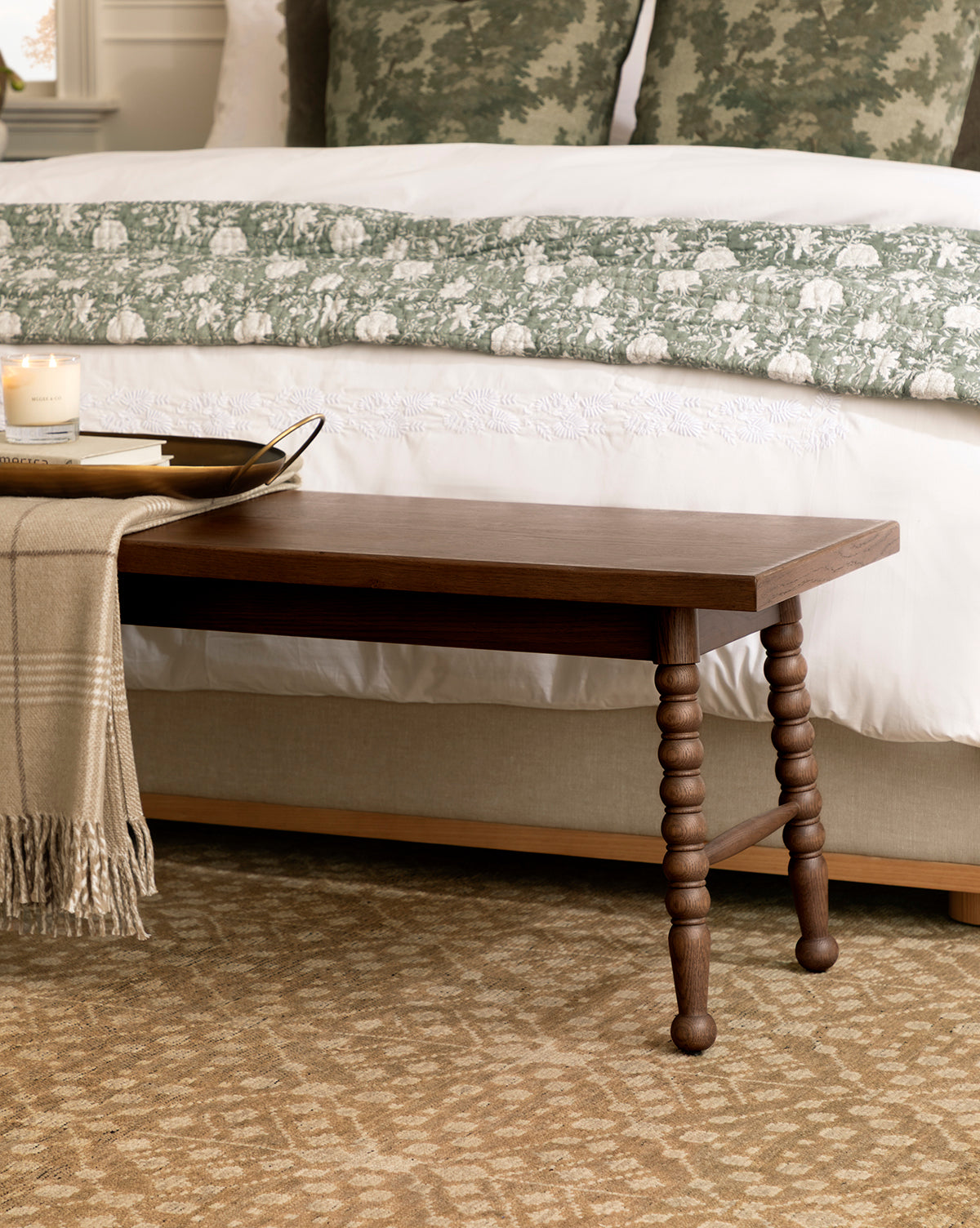 A Heathcliff Bench sits at the foot of a bed with green and white bedding; a tray holding a candle and book, along with a beige throw blanket, are arranged on the bench, by McGee & Co.