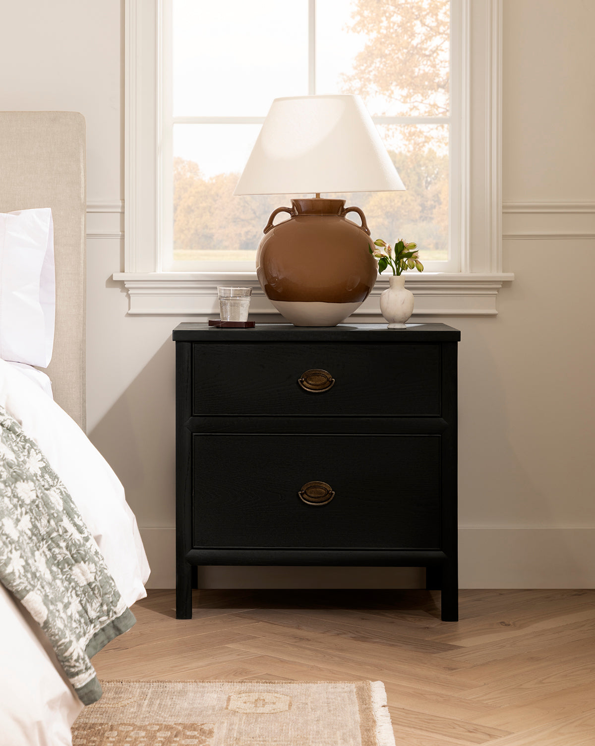 The Edward Nightstand in black ash with brass handles stands by the bed, topped with a brown vase lamp, a small plant, and a glass of water. Natural light from the window brightens this cozy bedroom setup by McGee & Co.