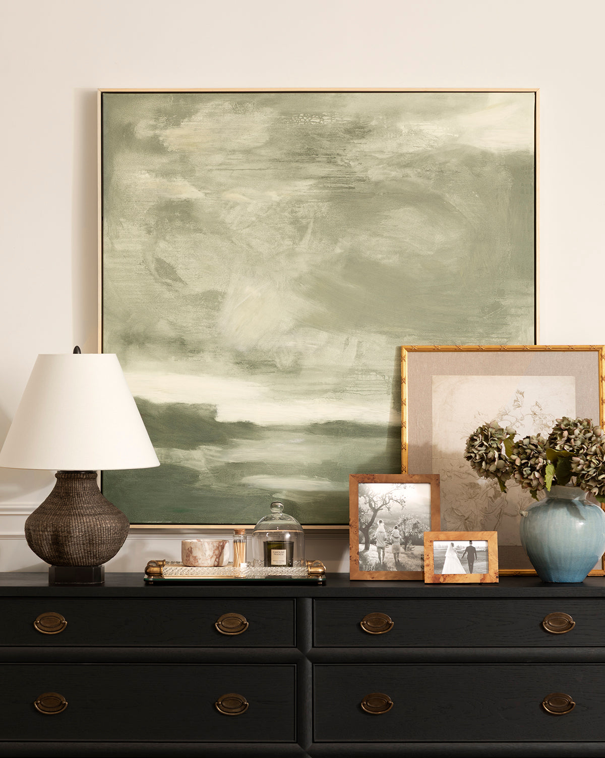 A dresser with round brass handles holds a textured lamp, a tray with a candle and framed photo, a blue vase featuring Feathery Floral, and grouped framed photos. Above hangs a large abstract green and white giclée on paper, by McGee & Co.