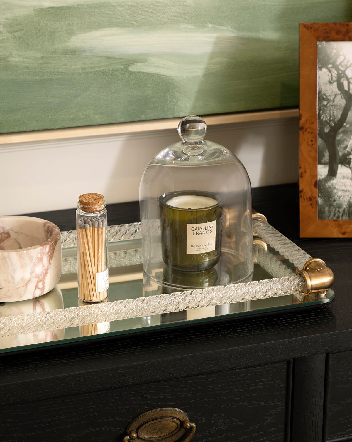 A glass cloche covers a green candle on a mirrored tray beside the Verona Marble Bowl, which has brown veining and a matte finish. A glass jar of matches, a framed black-and-white photo, and a green painting complete the elegant scene by McGee & Co.