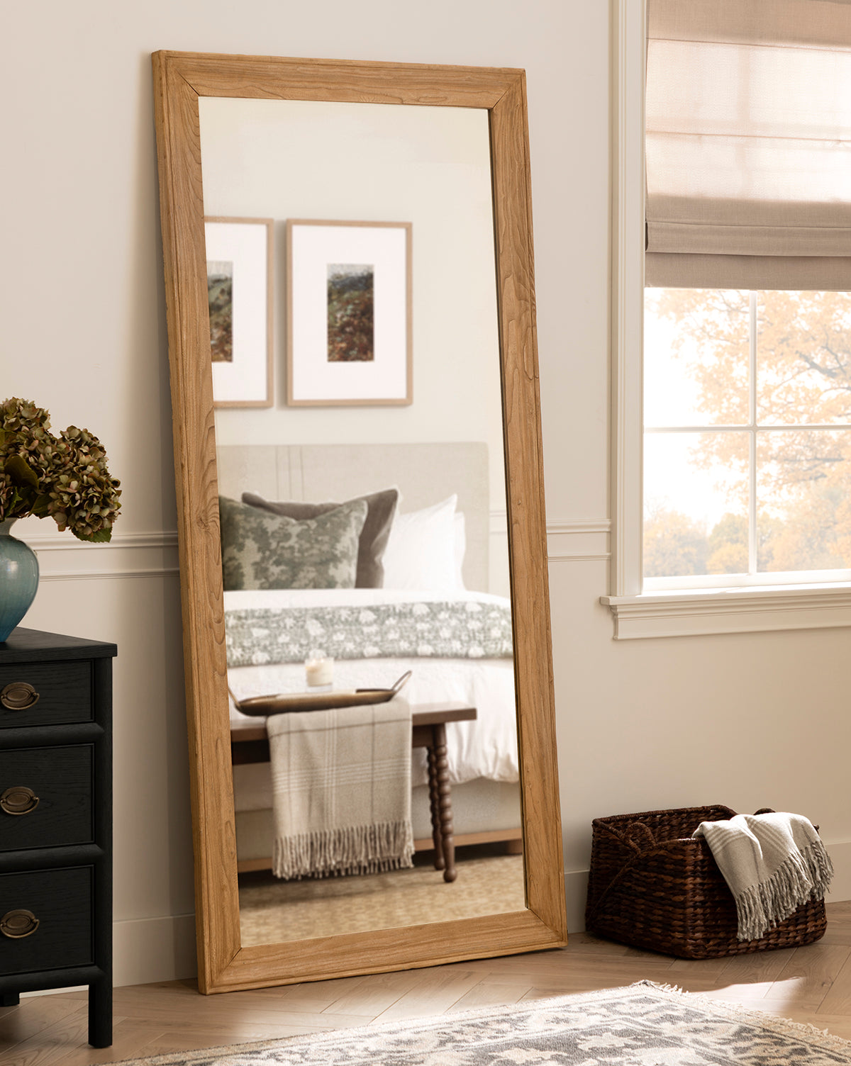 The Pippen Floor Mirror, featuring a honey elm finish, leans against the wall and reflects a bed with green and white bedding, a bench with a striped throw, two framed pictures, plus a nearby black dresser and window with beige blinds by McGee & Co.