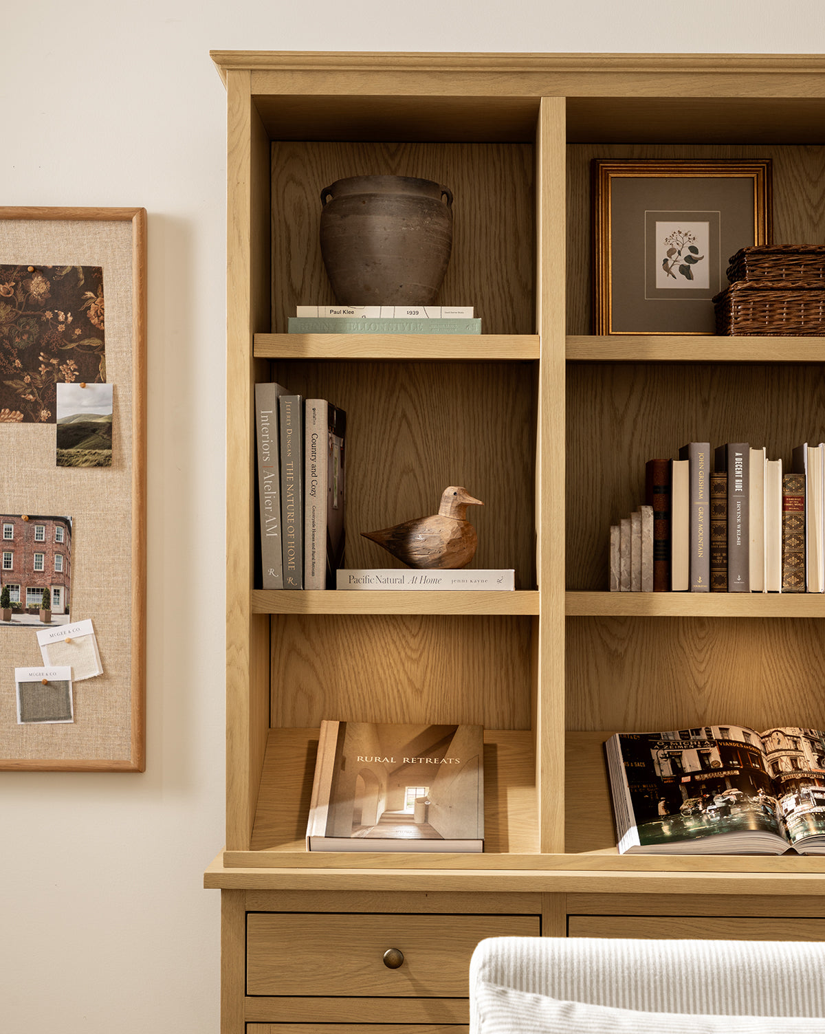 A wooden bookshelf displays books, a ceramic vase, a small framed artwork, a wicker basket, and the Wooden Bird Object to add nature-inspired charm. A bulletin board with photos and notes hangs on the wall to the left, by McGee & Co.