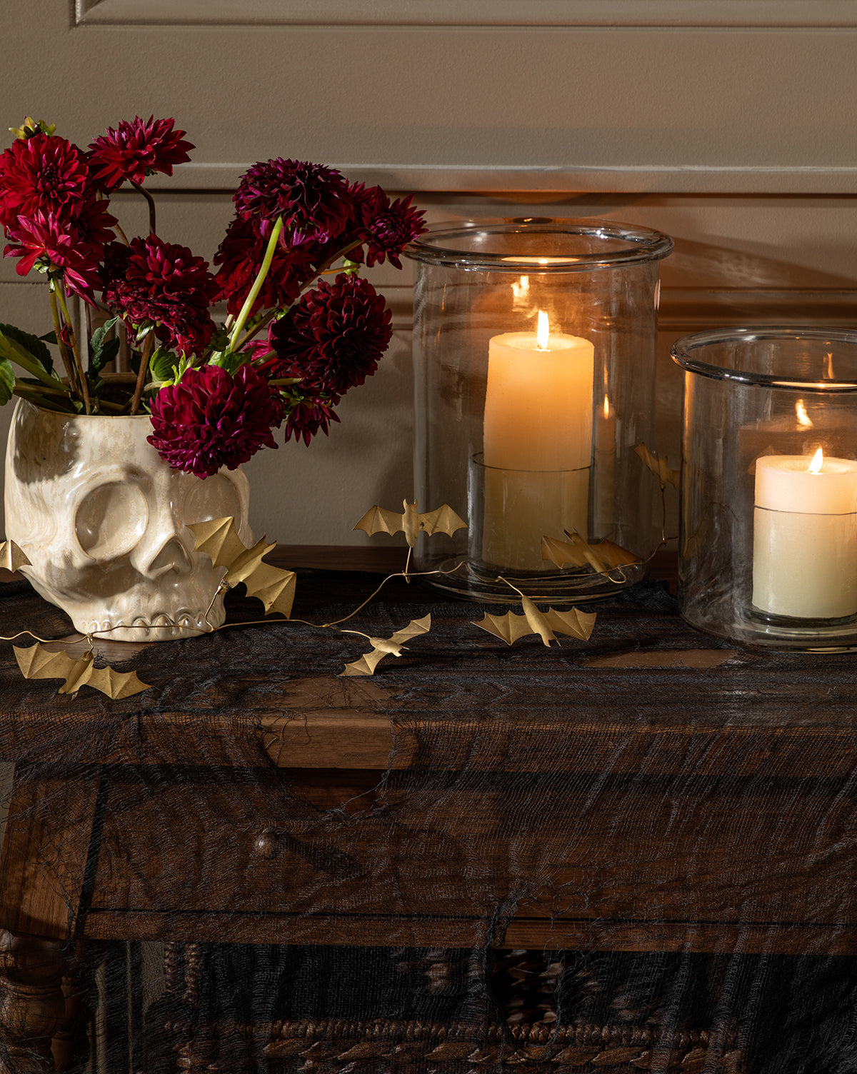 A skull-shaped vase with red flowers sits on a wooden table by two lit candles in glass holders. Halloween décor includes dark fabric, paper bats, and the Gold Metal Bats Garland 72 for a striking, golden autumnal accent by McGee & Co.