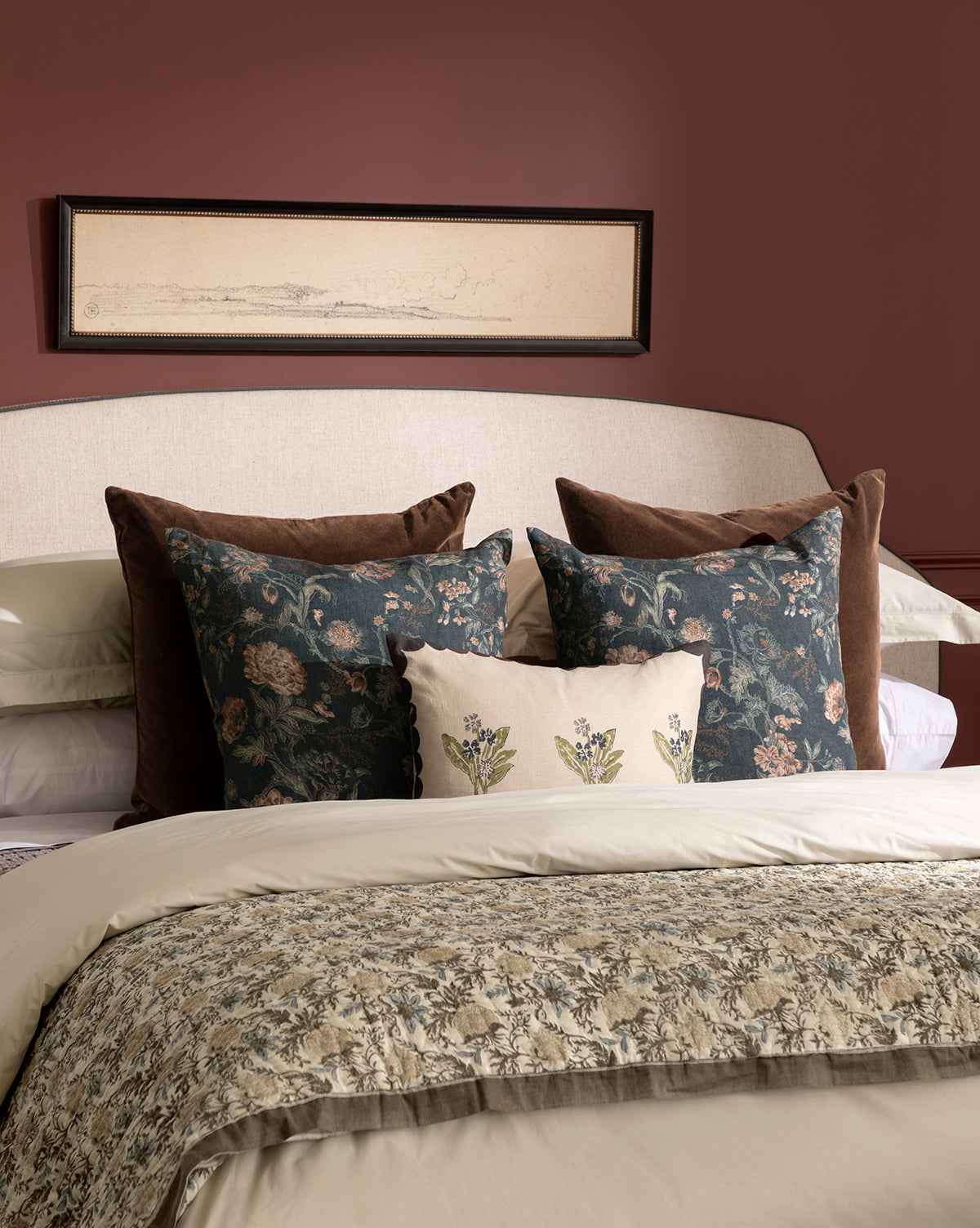 A neatly made bed dressed in Clouded Island bedding with cream and patterned blankets and layered floral embroidered pillows, set before a padded headboard and mauve wall featuring a black-framed landscape sketch above, by McGee & Co.
