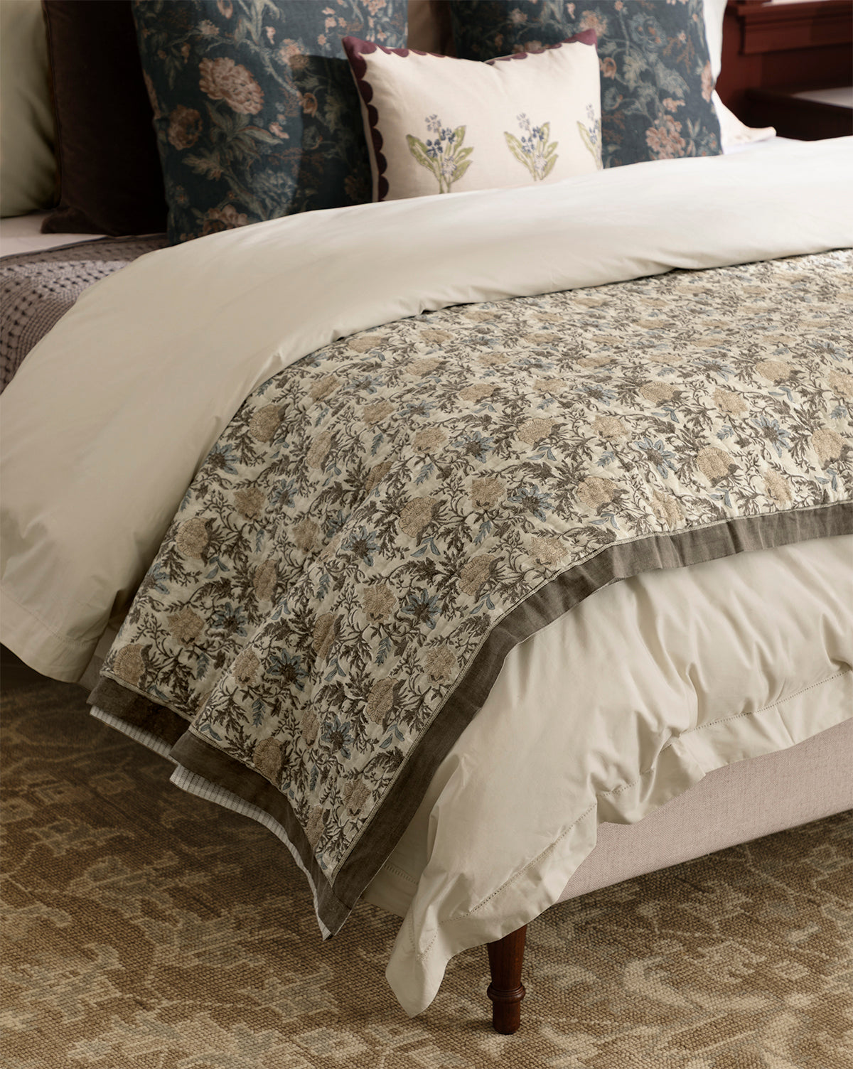 A cozy bedroom features a neatly made bed with cream-colored bedding, the Audrey Tan Block Print Quilt with ruffle edging, and floral decorative pillows, set atop a patterned carpet, by McGee & Co.