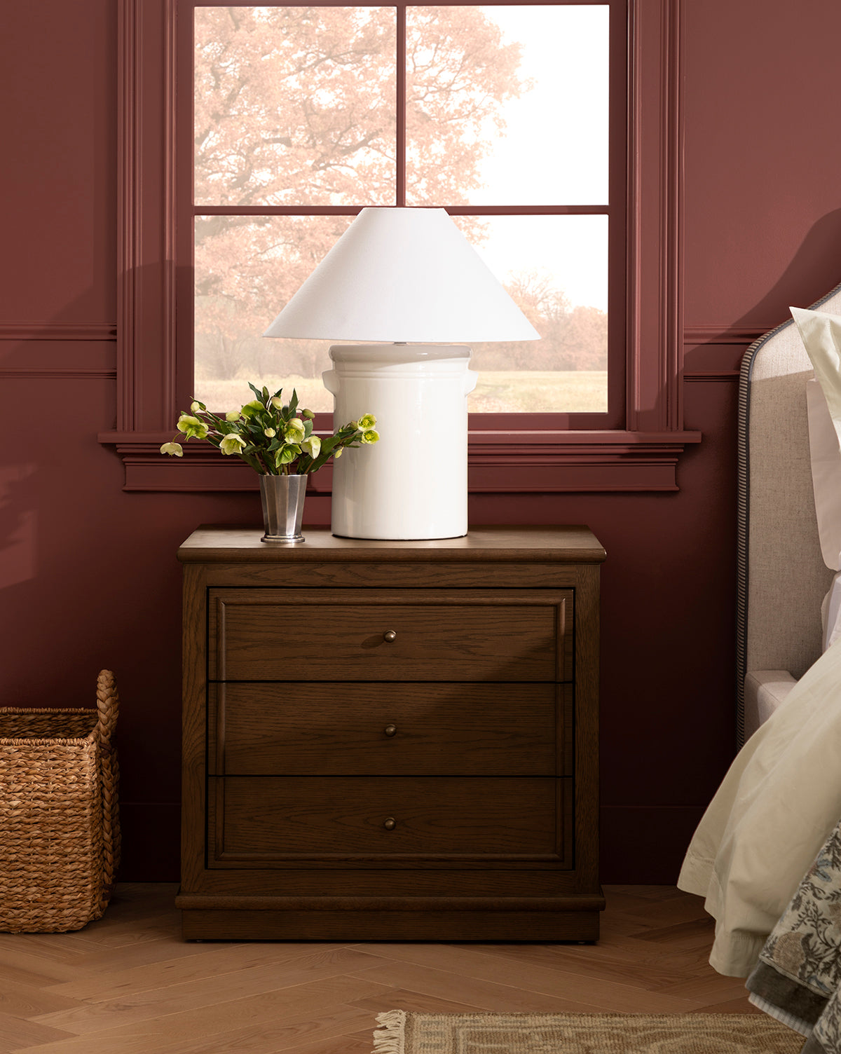 A Yorkshire Nightstand with an English oak finish and antique brass pulls stands by the bed, topped with a white lamp and vase of yellow flowers. Pinkish-red window trim frames leafy trees, and a wicker basket sits on the floor nearby, by McGee & Co.