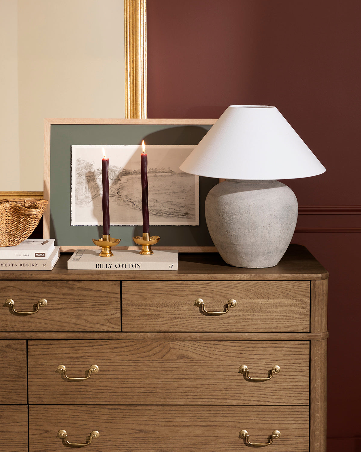 A wooden dresser with antique brass handles holds a white lamp, elegant decor like dark red candles in Cosima Taper Holders, stacked books, a framed picture, and a basket, all set against a maroon wall with gold-framed artwork by McGee & Co.