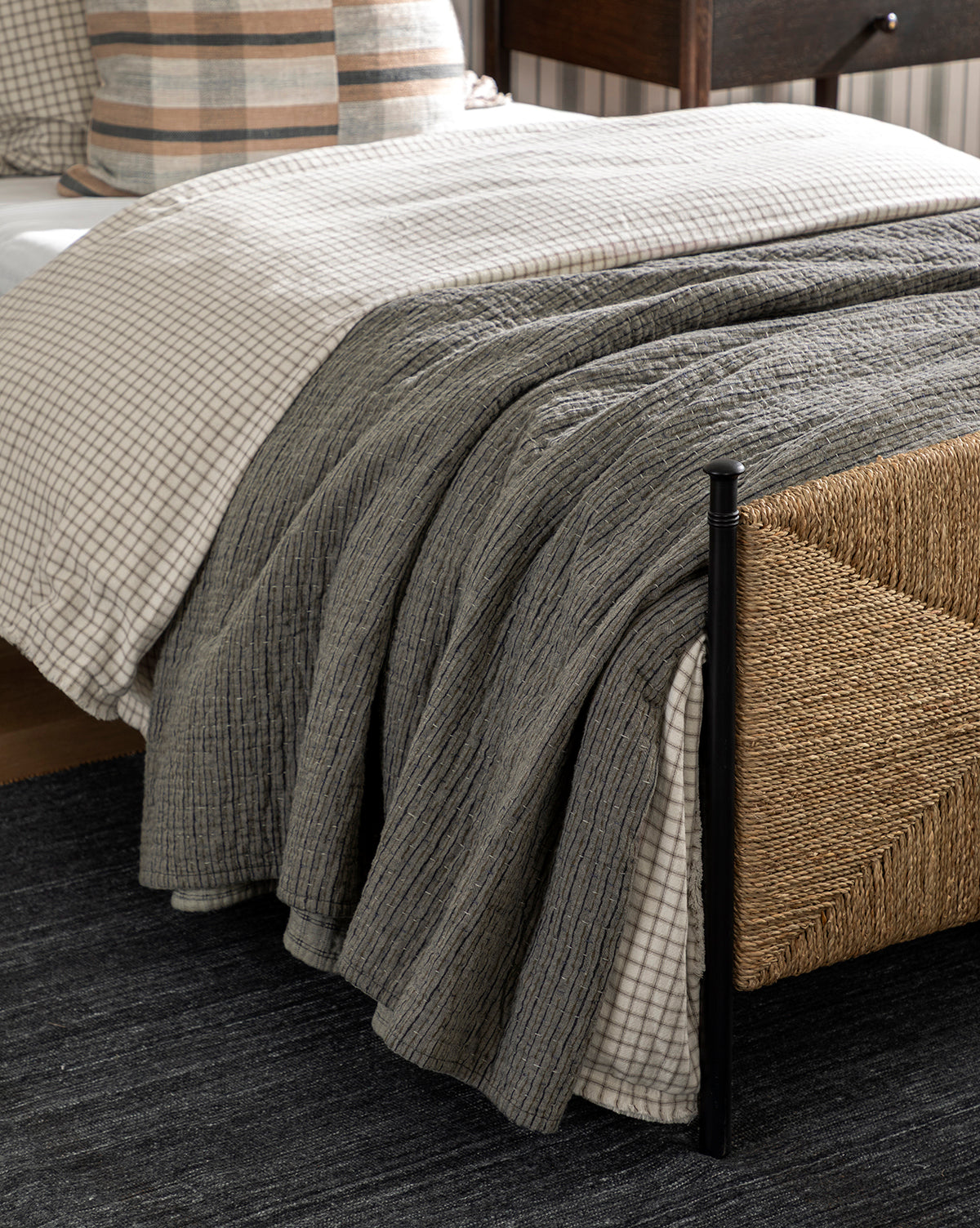 A cozy bed with a checkered duvet, textured gray blanket, and plaid pillows features the charming Cecily Kids Quilt. A woven rattan footboard and a dark rug enhance the warm, inviting, rustic vibe by McGee & Co.
