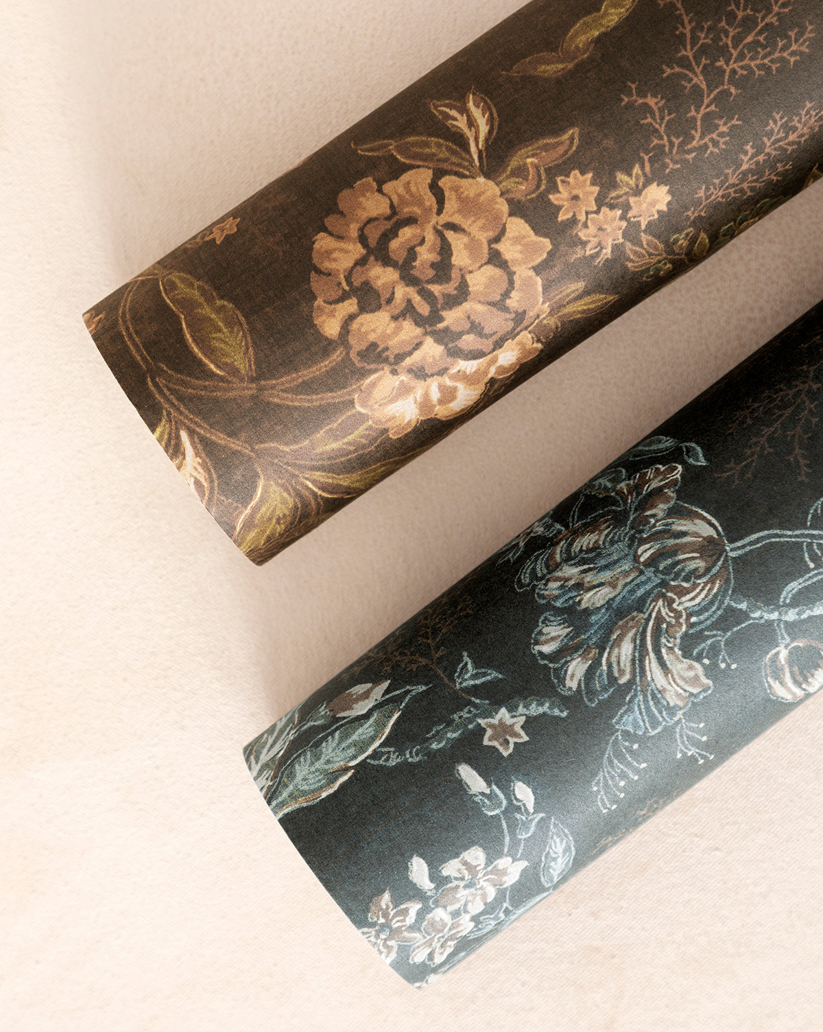 Two rolls of Hestercombe Floral Wallpaper—one with gold and brown flowers, the other with blue and green—are displayed on a light beige surface by McGee & Co.