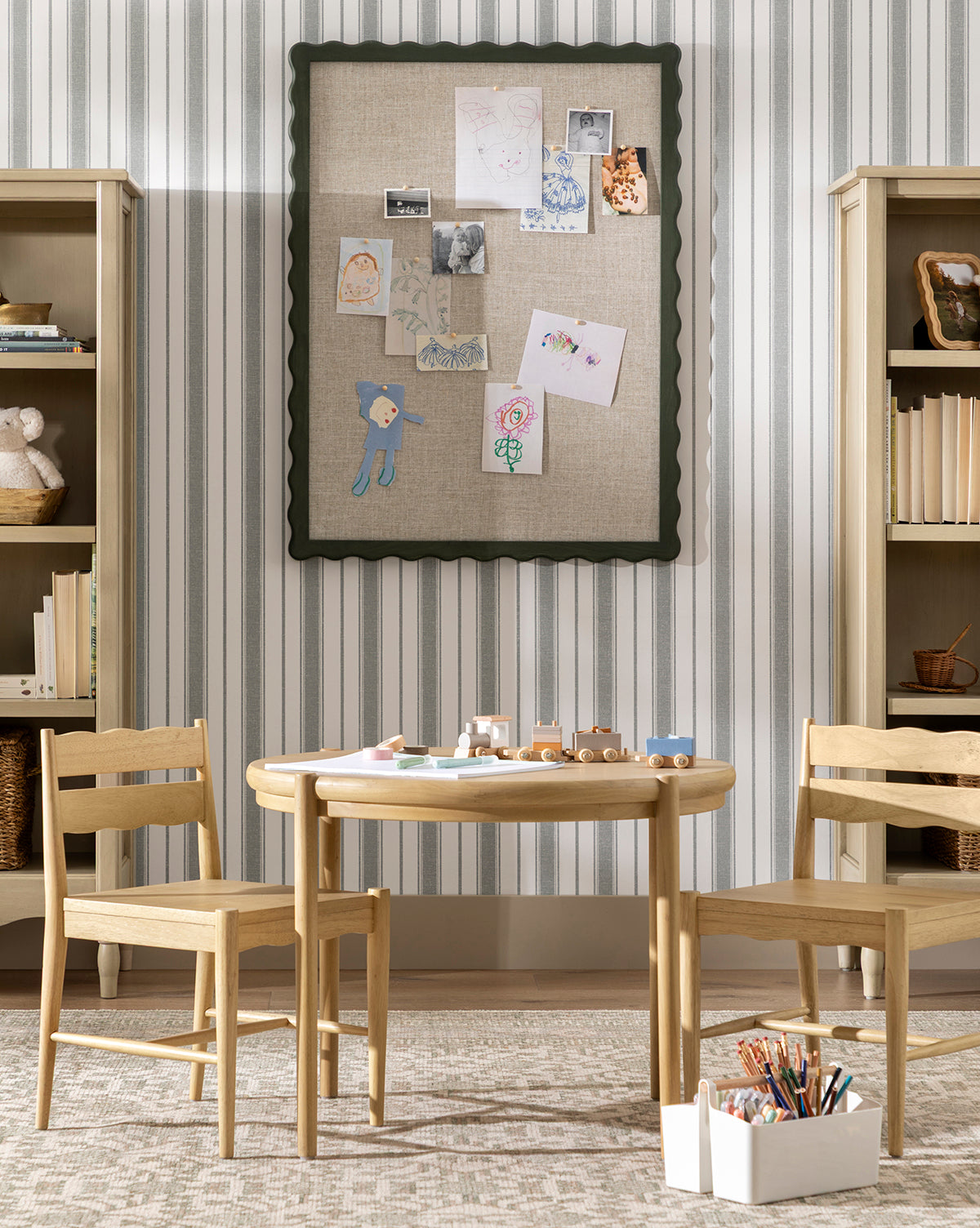 A children’s playroom features a round white oak Darcy Kids Table with two chairs, art supplies, and toy cars. Behind it, a bulletin board displays drawings and photos, flanked by bookshelves on striped wallpaper, by McGee & Co.