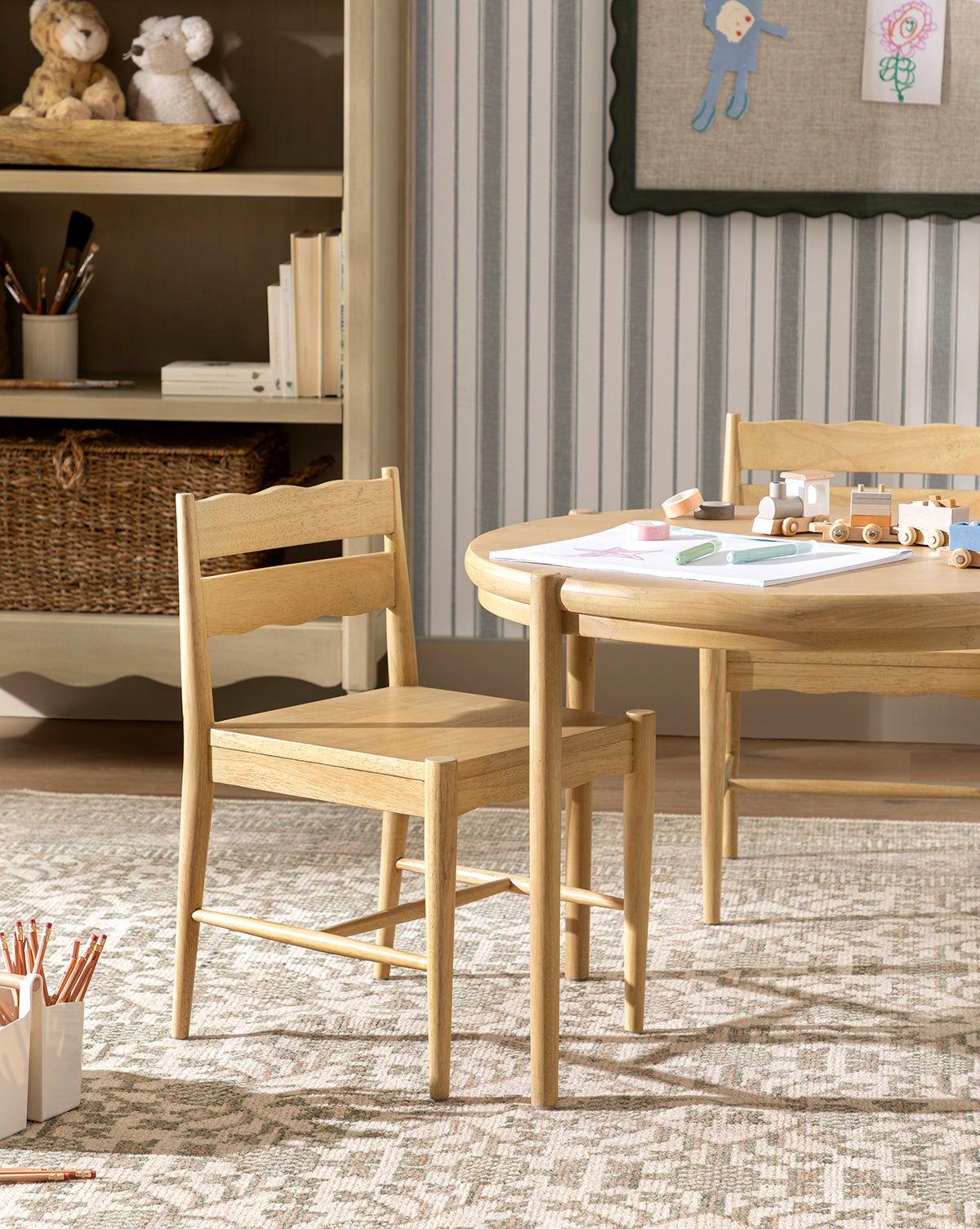 Two Darcy Kids Chairs in light white oak and a round table with art supplies sit on a patterned rug in a cozy room, with shelves holding baskets, books, and stuffed animals in the background by McGee & Co.