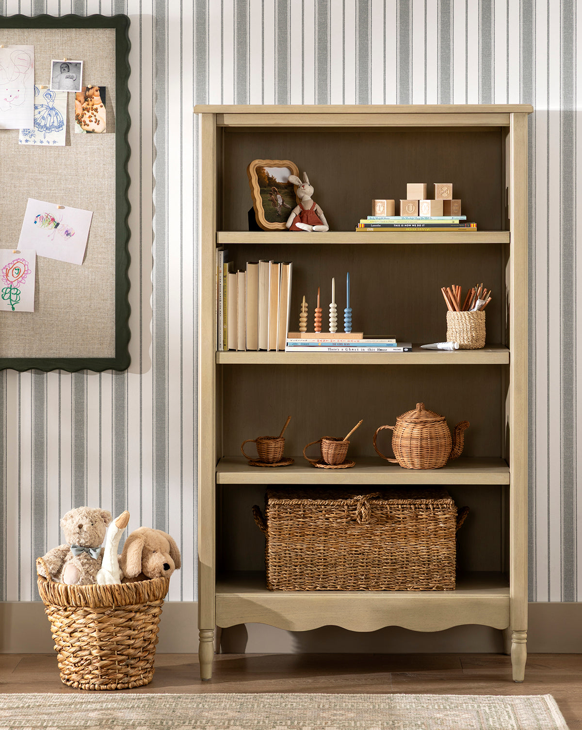 The Liberty Bookcase stands against a striped wall, displaying books, a toy rabbit, wooden stacking toys, cups, and baskets. Childrens drawings are pinned to a corkboard on the left, by McGee & Co.