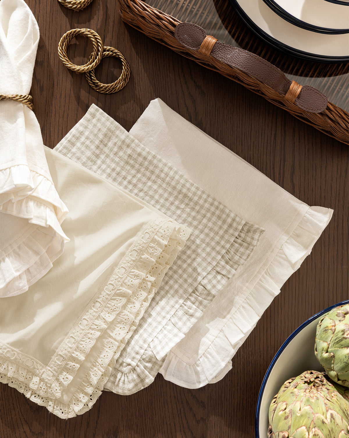 A tabletop displays Catania Linen Napkins (Set of 4) with assorted patterns and ruffled or lace trim, beside a woven tray, stacked plates, gold napkin rings, and a bowl of artichokes by McGee & Co.