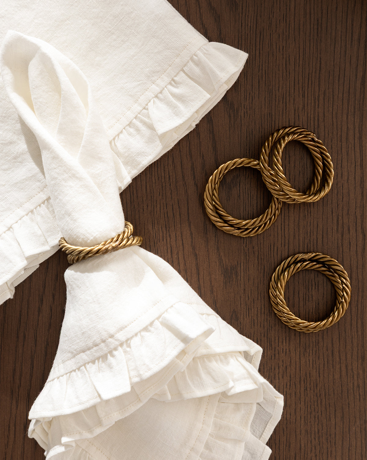 A folded white Catania Linen Napkin with ruffled edges is held by a gold twisted napkin ring, with three more gold napkin rings beside it on a wooden surface, by McGee & Co.