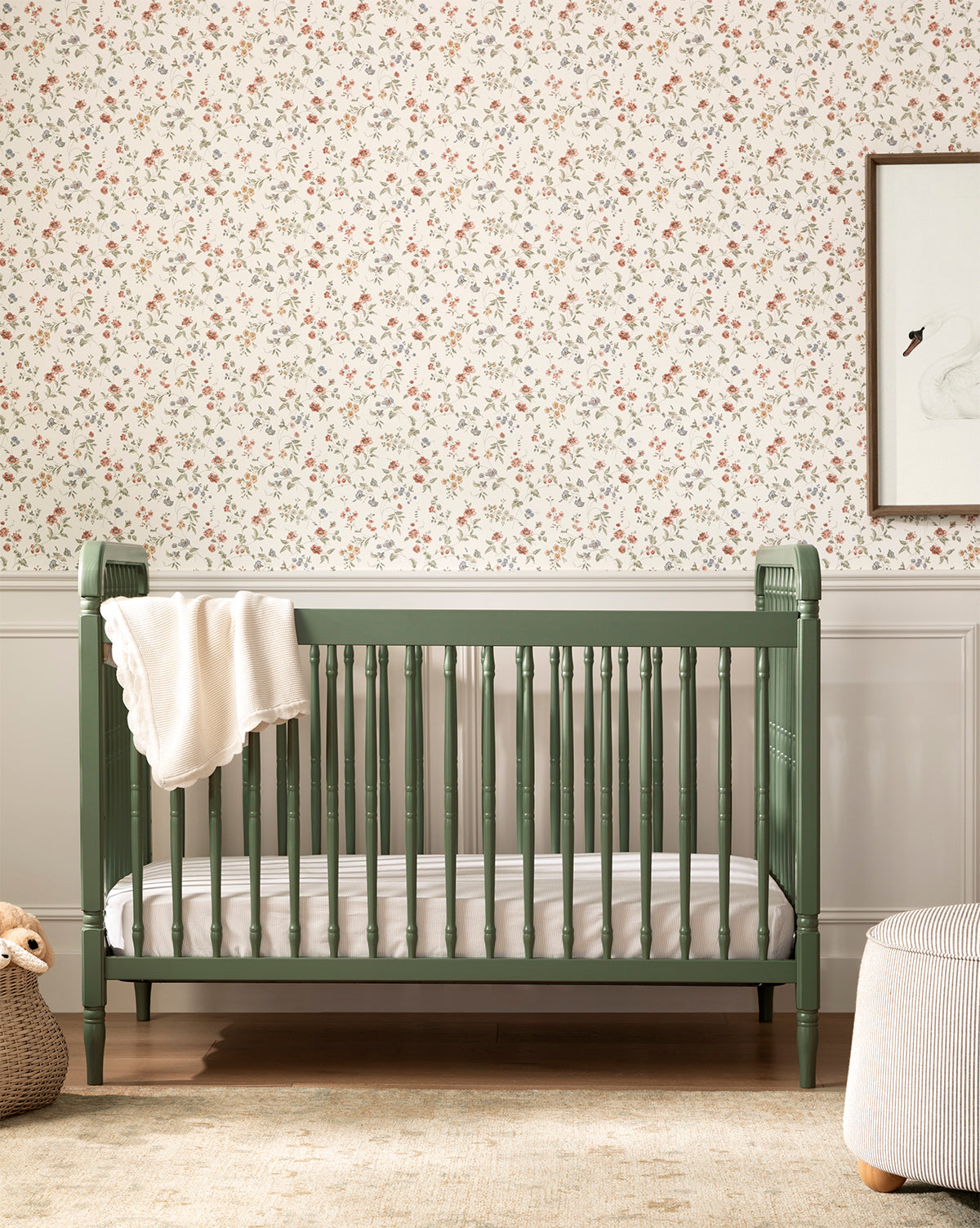 The Liberty 3-in-1 Convertible Spindle Crib w/Toddler Bed Conversion Kit in green, made from solid pine wood, stands before floral wallpaper with a white blanket draped over the side, by McGee & Co. A woven basket with a plush toy is nearby.