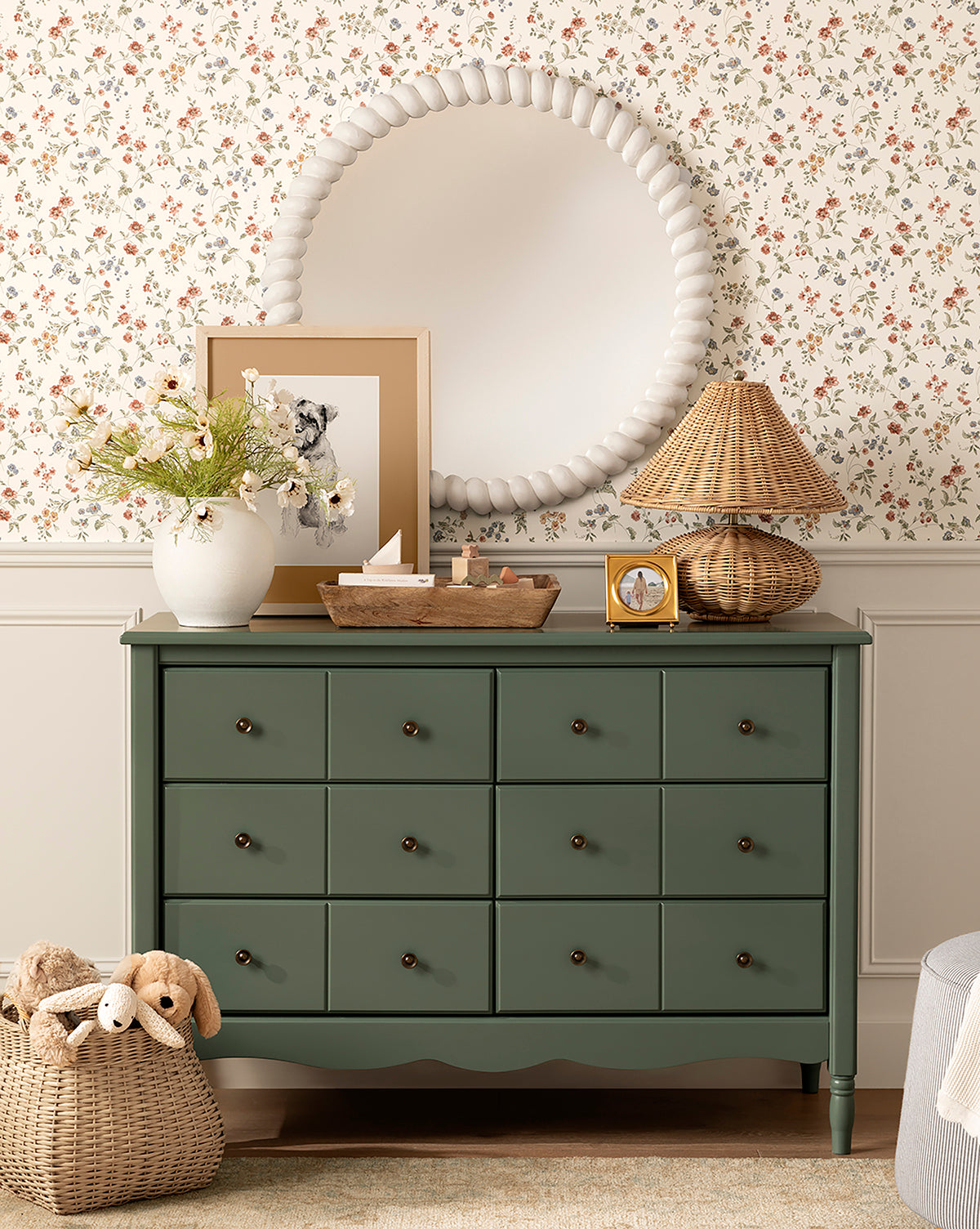 The Liberty 6-Drawer Assembled Dresser, made from sustainable pine with brass knobs, stands against floral wallpaper and is styled with a beaded mirror, framed art, wicker lamp, vase of flowers, wooden tray, and a basket of stuffed animals on the floor, by McGee & Co.