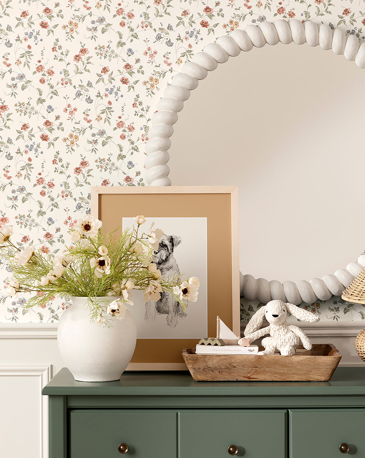 A green dresser with a white vase of flowers, framed dog sketch, Chenille Bunnies (Set of 2), and a toy boat sits below a round beaded mirror. Floral wallpaper and white wainscoting complete this perfect nursery decor, by McGee & Co.