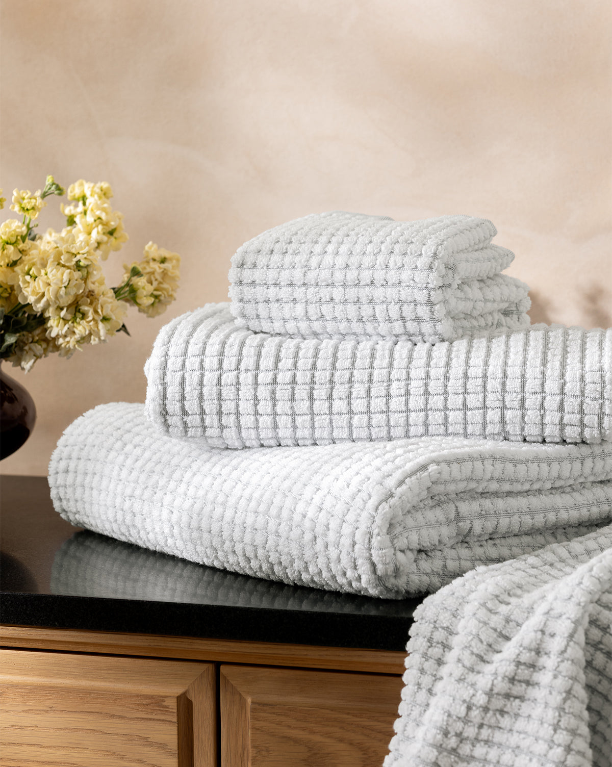 A neatly stacked set of Atrani Gray Organic Bath Collection waffle weave towels sits on a black countertop next to a vase of yellow flowers, against a light beige wall, by McGee & Co.