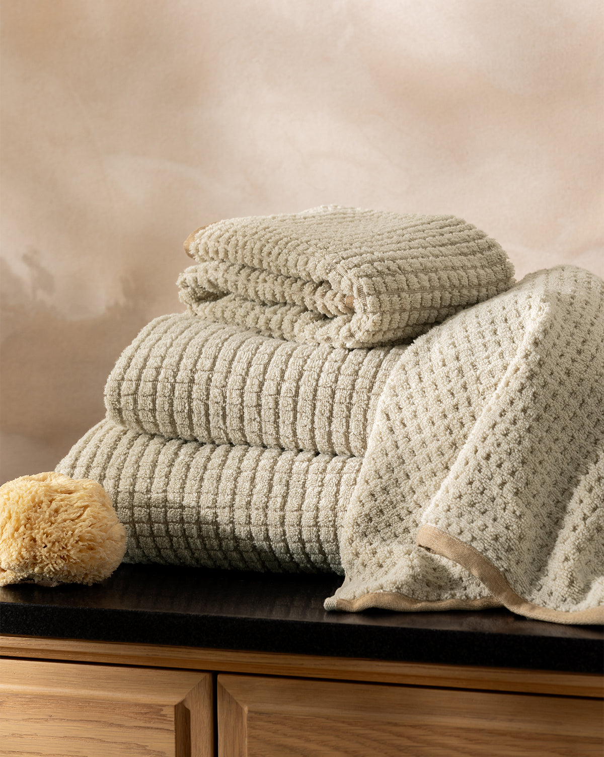 The Atrani Organic Bath Collection, featuring beige textured cotton towels folded and draped on a dark countertop beside a natural sponge, is set against light wood cabinets and a soft beige background, by McGee & Co.