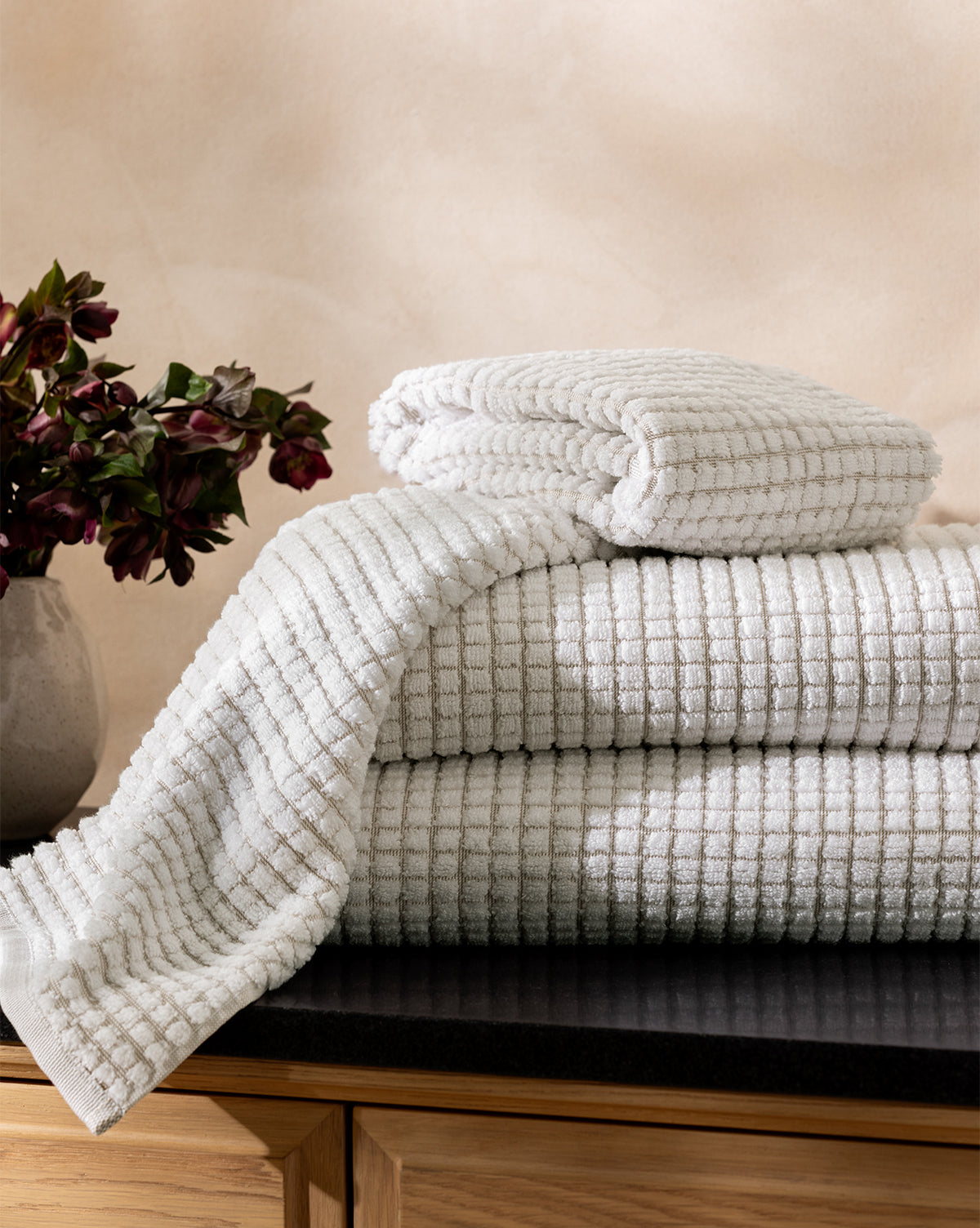 The Atrani Tan Organic Bath Collection, made of organic cotton with a waffle weave, is neatly folded on a dark countertop beside dark red flowers, set against light walls and wooden cabinets, by McGee & Co.
