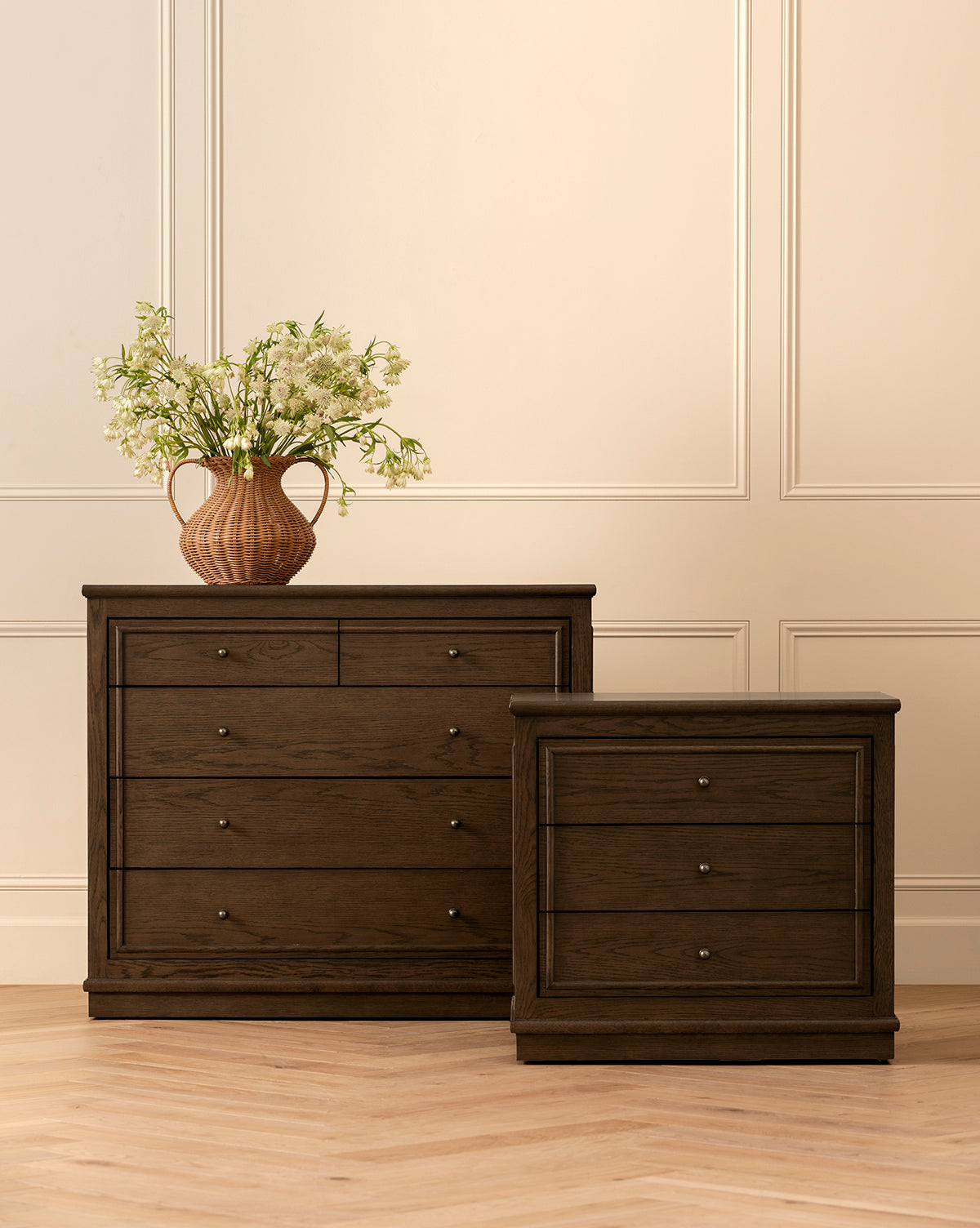 Two Yorkshire Nightstands with antique brass pulls, one taller and one shorter, stand side by side on a light herringbone floor. The taller nightstand holds a brown vase with white flowers against a cream-colored paneled wall, by McGee & Co.