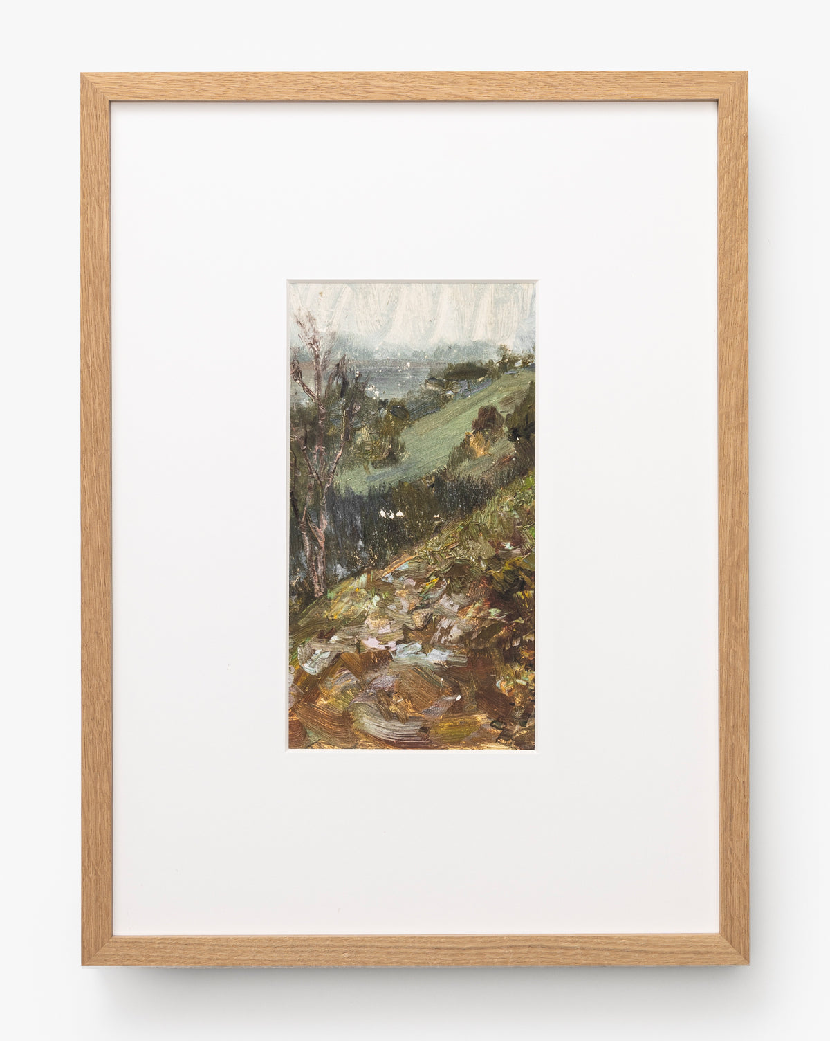 A framed painting titled A River Runs Through features a small, vertical landscape of grassy hills, trees, and rocks beneath a cloudy sky in earthy green and brown tones with a light wood frame and white mat, by McGee & Co.