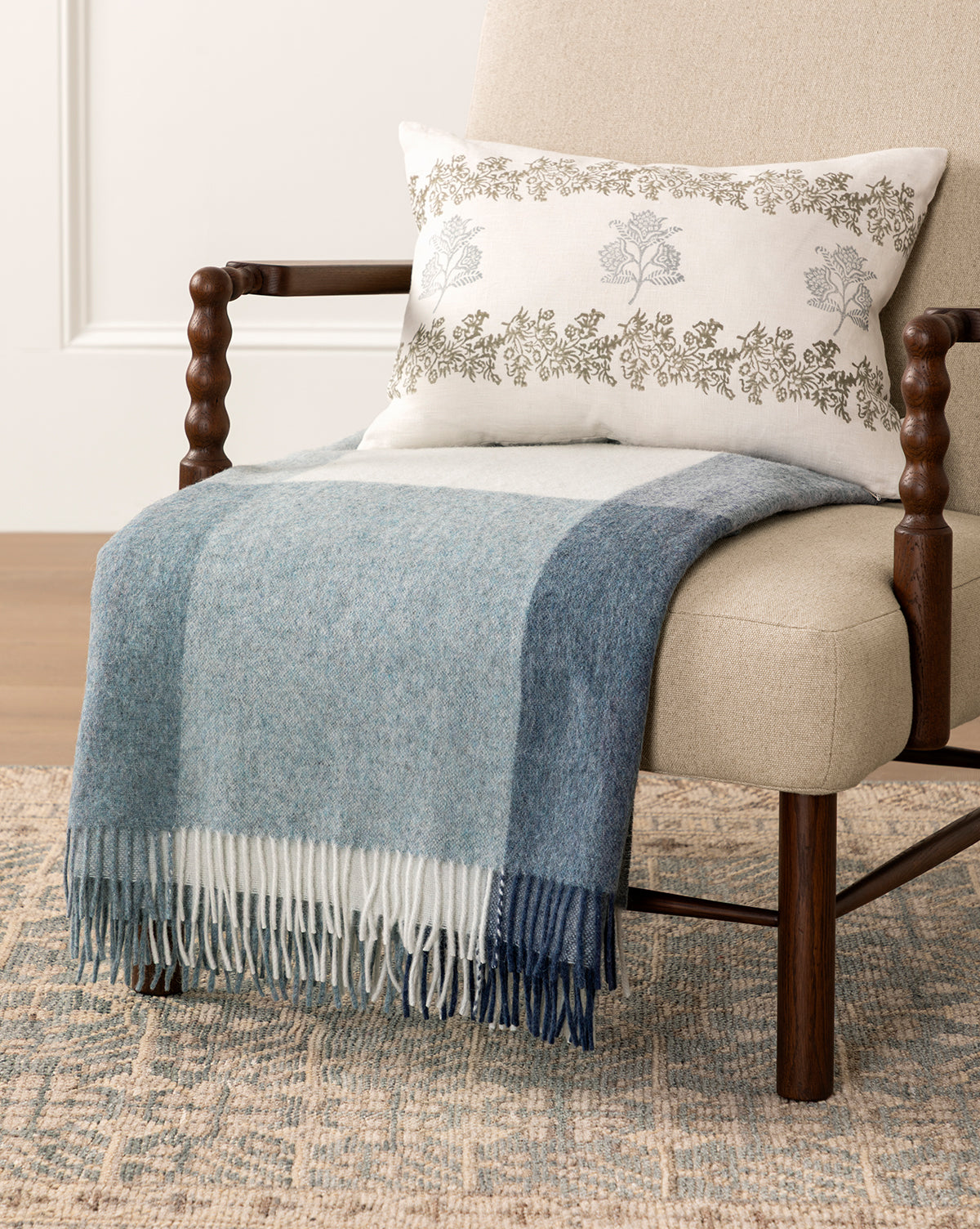 A beige armchair with turned wooden arms is draped with the eco-friendly Abner Throw in blue and gray stripes, and topped with a white pillow featuring floral embroidery, all set on a textured rug, by McGee & Co.