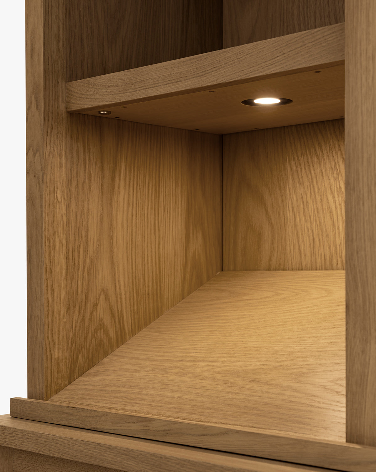 A close-up of the Adler Oak Cabinet shows its wooden shelf with a slanted back panel and integrated lighting from a small, built-in round LED under the upper shelf, by McGee & Co. The cabinet features a natural, light oak finish.