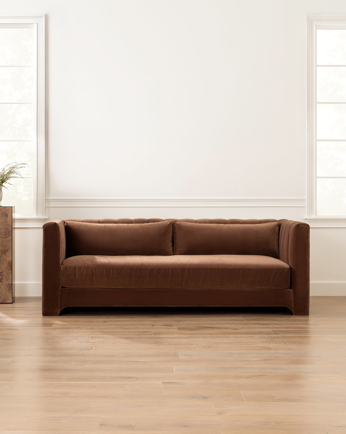 A modern living room showcases the Agatha Sofa 90 in brown velvet against a white wall, bordered by tall windows, by McGee & Co.