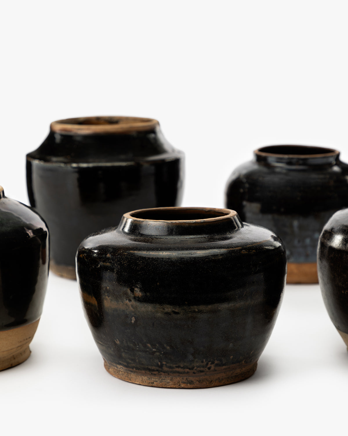 Four Agnes Vases with wide bodies and narrow openings are arranged against a white background. The black-glazed ceramic pottery has a rustic, handmade look with earthy brown bases, by McGee & Co.