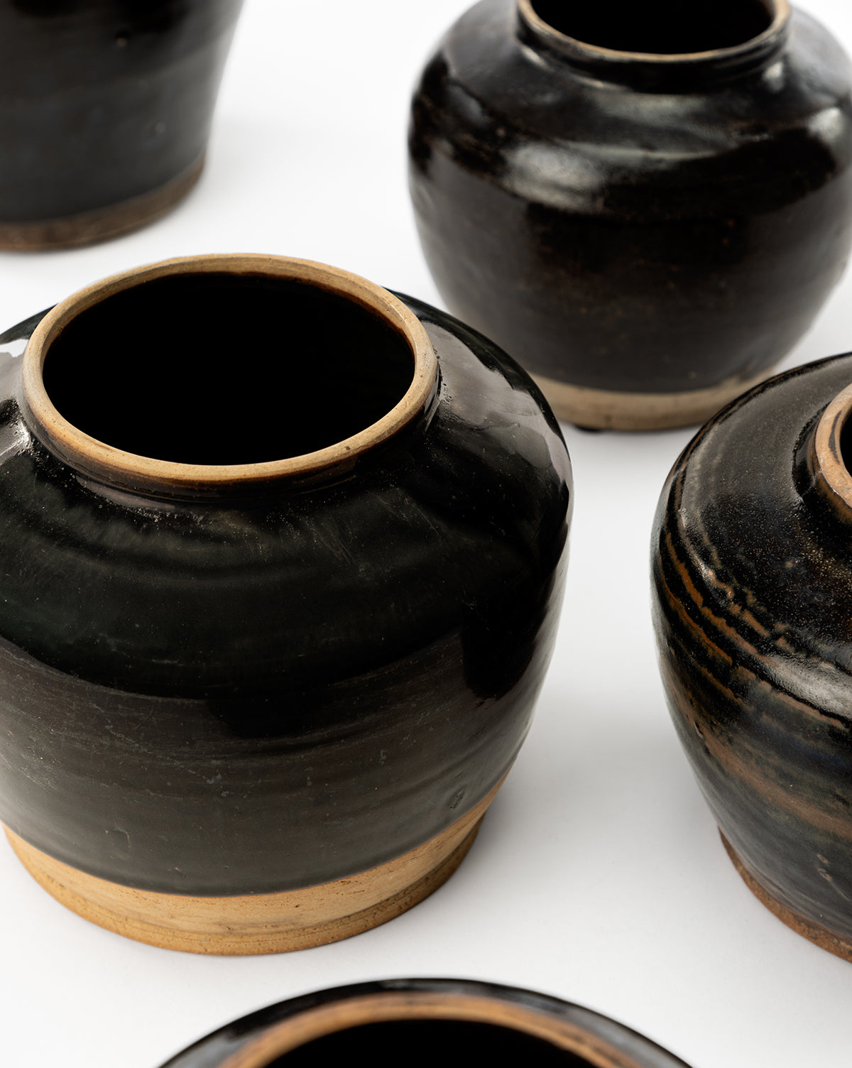Several Agnes Vases, round and black with wide openings and unglazed light brown bases, are arranged on a white surface. These ceramic vases have a glossy finish and are positioned at various angles, by McGee & Co.
