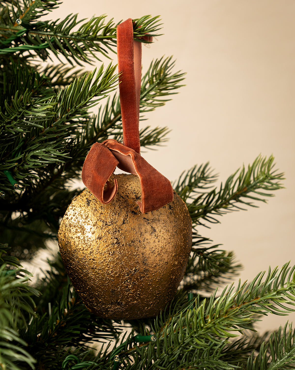 The Antique Gold Metal Bell with Velvet Ribbon hangs on a green Christmas tree branch, adding elegant holiday charm to your décor with its antique finish and luxurious brown velvet ribbon, by McGee & Co.