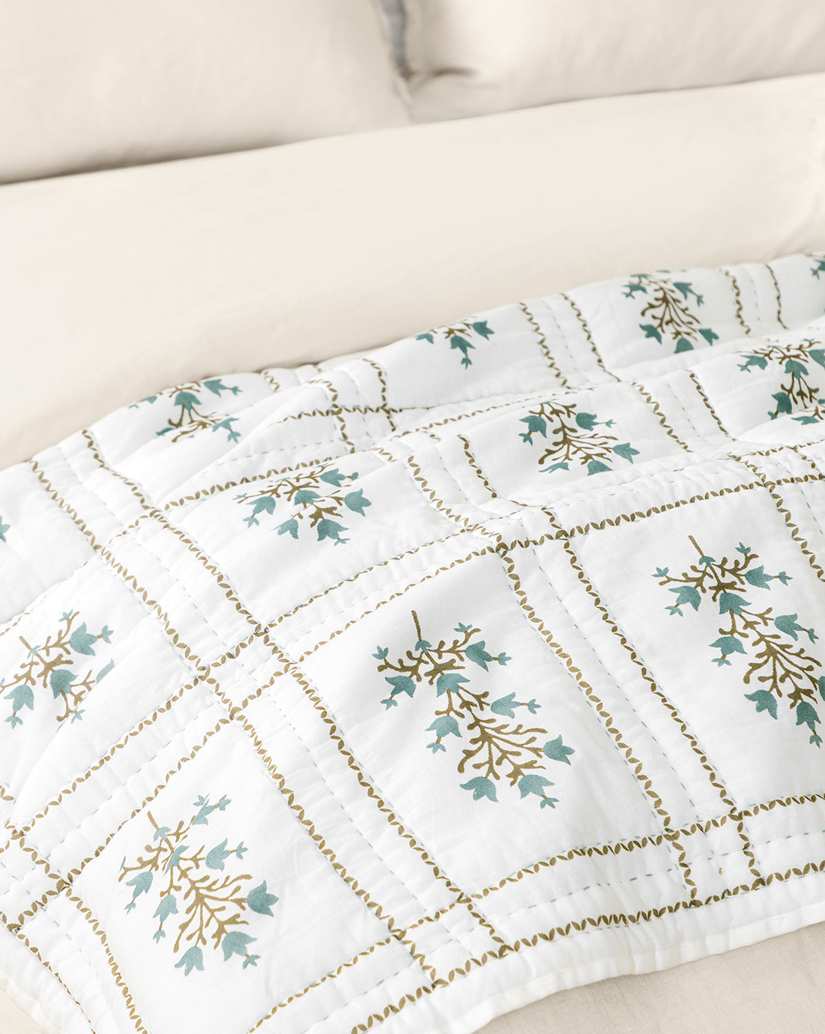 The Atherton Quilt, featuring a white floral design with blue and gold accents and a stitched grid pattern, highlights handmade artistry on a cream bed with matching pillows, by McGee & Co.