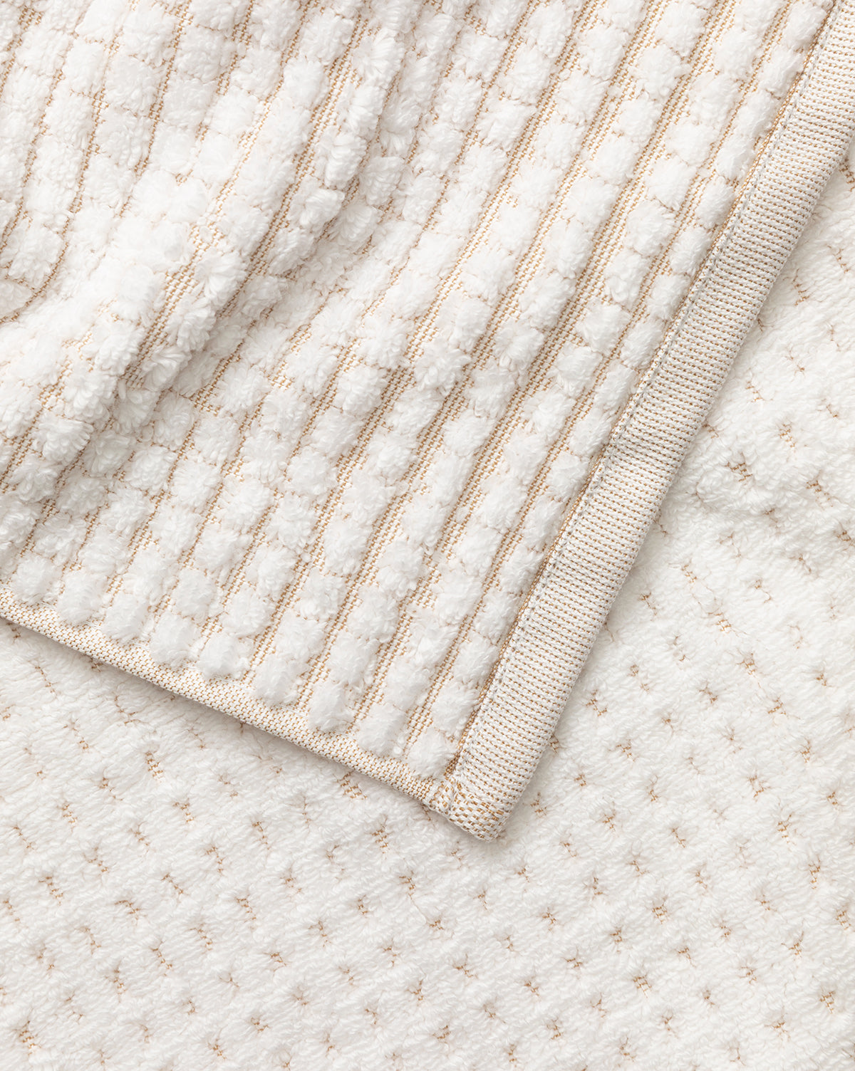 A close-up of a folded white waffle weave towel from our Atrani Tan Organic Bath Collection, featuring a textured striped pattern and beige trim, displayed on a subtly dotted white surface by McGee & Co.
