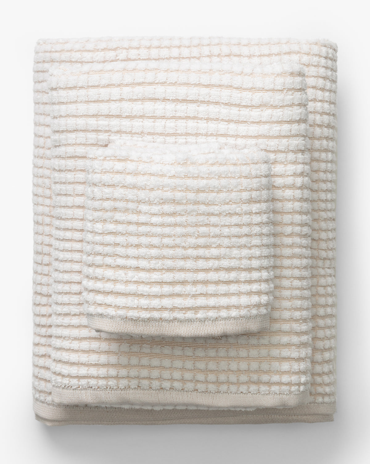 A neatly folded stack of three cream and beige striped towels in varying sizes from the Atrani Tan Organic Bath Collection, arranged largest to smallest on a white background, by McGee & Co.
