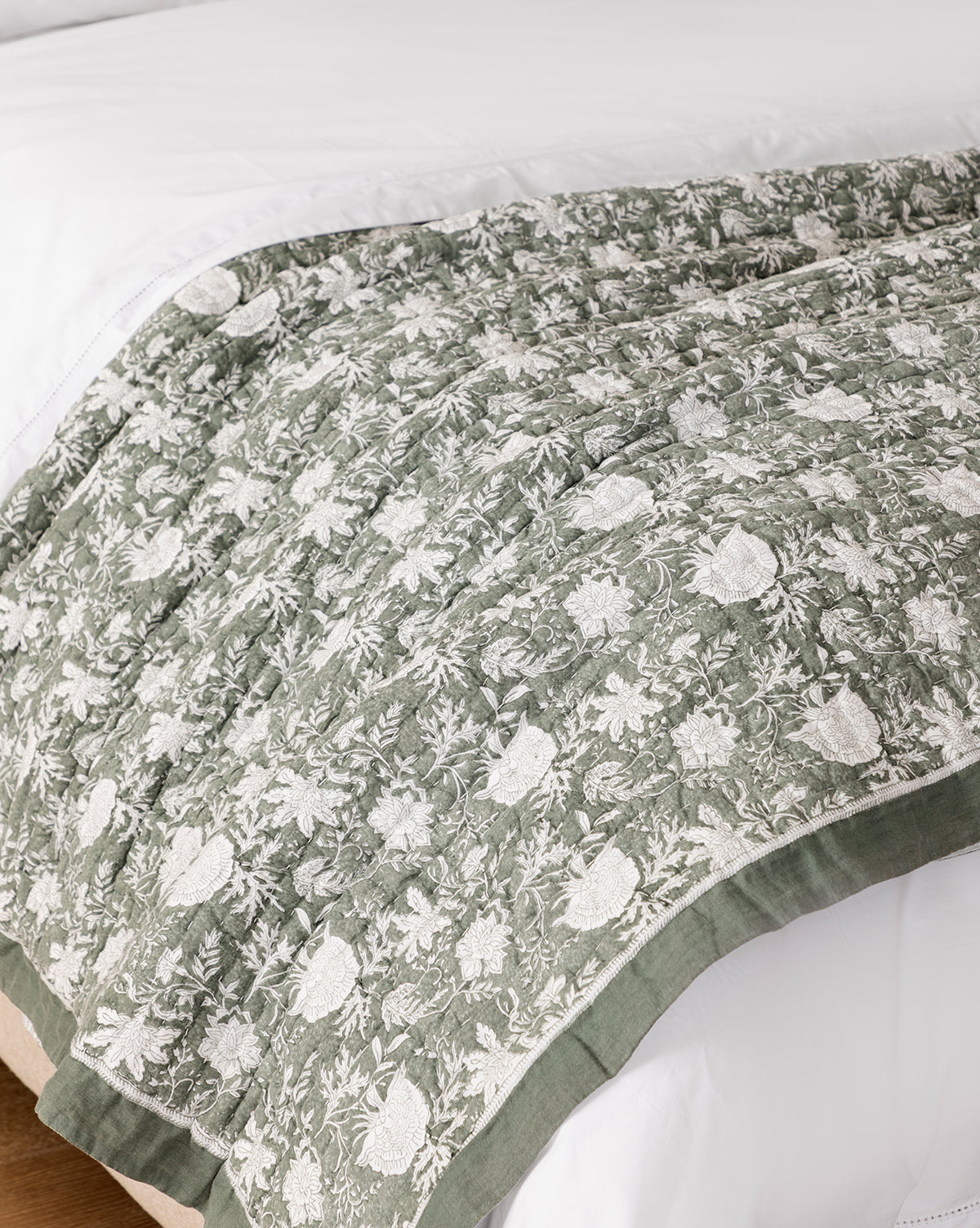 The Audrey Sage Block Print Quilt, featuring an intricate floral pattern and decorative border, is draped across the foot of a white bed, partially covering a white comforter and resting gracefully on a wooden floor, by McGee & Co.